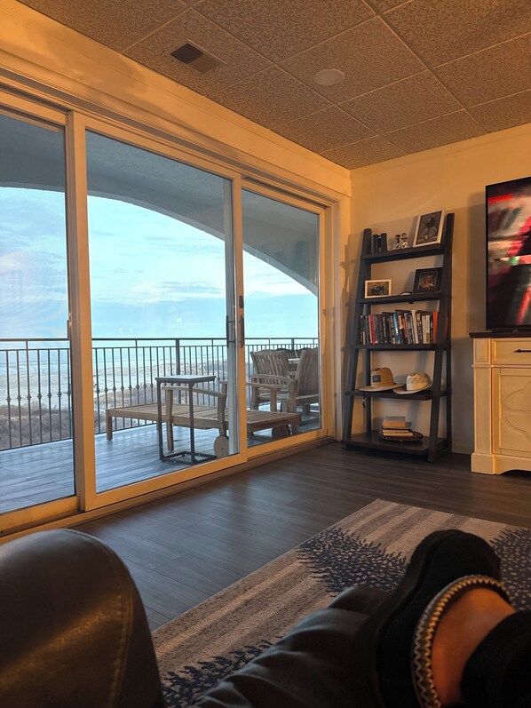 A living room with a large sliding glass door leading to a balcony with outdoor furniture. There is a bookshelf with books and decorative items, and a television on the right side.