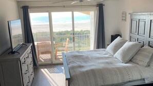 Een slaapkamer met een bed, een dressoir, een televisie en uitzicht op het strand door een schuifdeur.