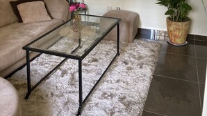 Un salon avec un canapé beige, une table basse en verre, un tél éviseur à écran plat fixé au mur, une plante en pot et un tapis blanc moelleux.