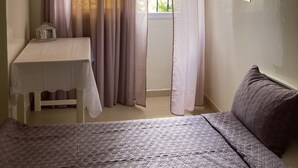 Une chambre à coucher avec un lit, une petite table, une fenêtre avec des rideaux et un appareil de climatisation.