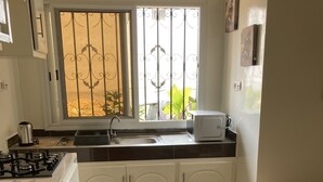 Une cuisine compacte équipée d’une cuisinière à gaz, d’un évier et de meubles blancs. Une fenêtre ornée de barreaux en métal décoratifs laisse entrer la lumière naturelle, et un canapé beige se trouve dans l’espace salon.