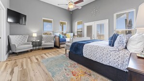 A modern bedroom with a large bed, two armchairs, a TV, and a ceiling fan.