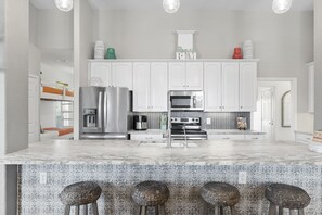 A modern kitchen with white cabinetry, stainless steel appliances, a marble island with bar stools, and decorative items on the shelves. - Game Room-Beach Views-Incredible Breeze-Sleeps 14- Child Friendly (Bolivar Peninsula)