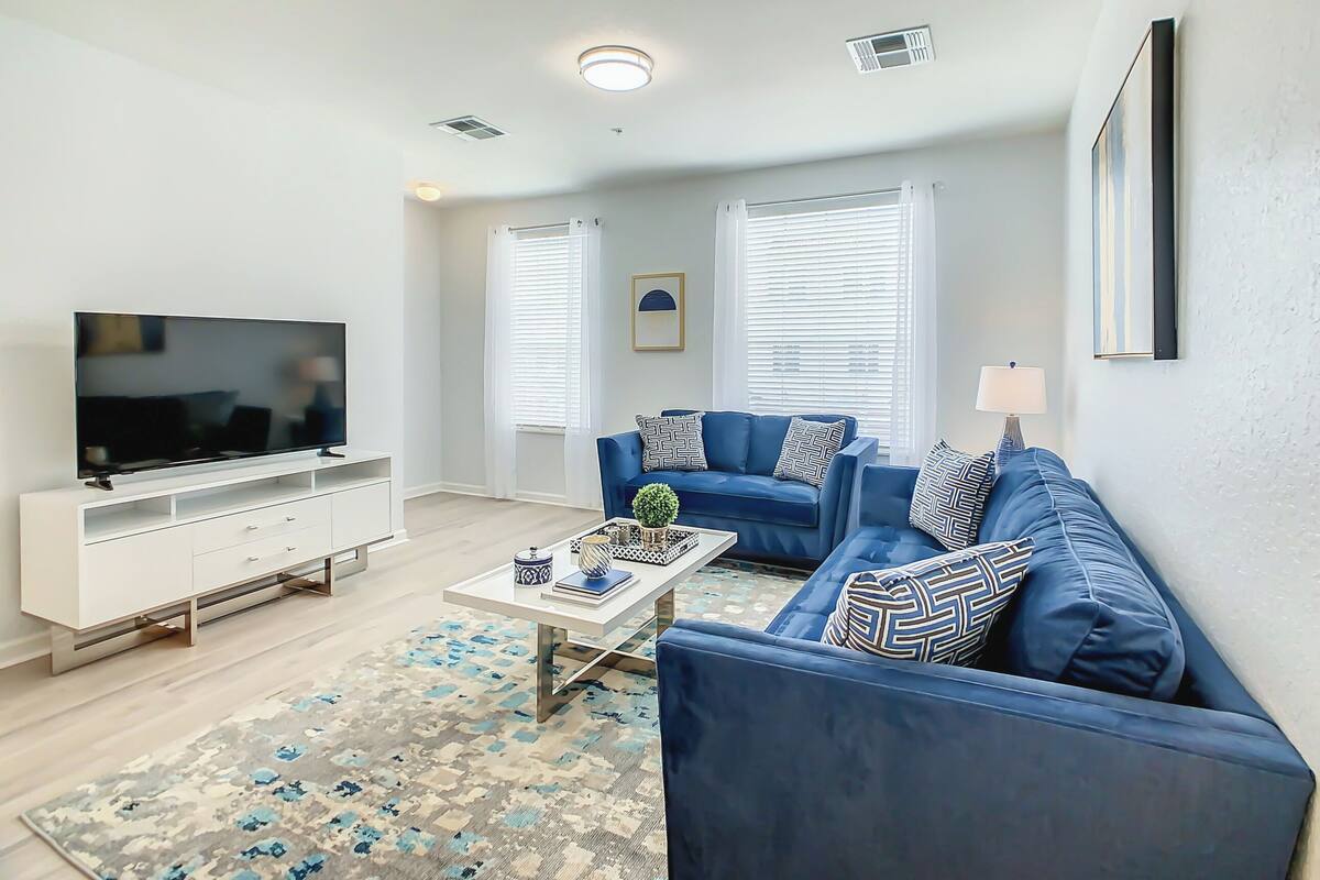 A modern living room with a flat-screen TV, a white entertainment unit, a blue sofa set, and a patterned rug.