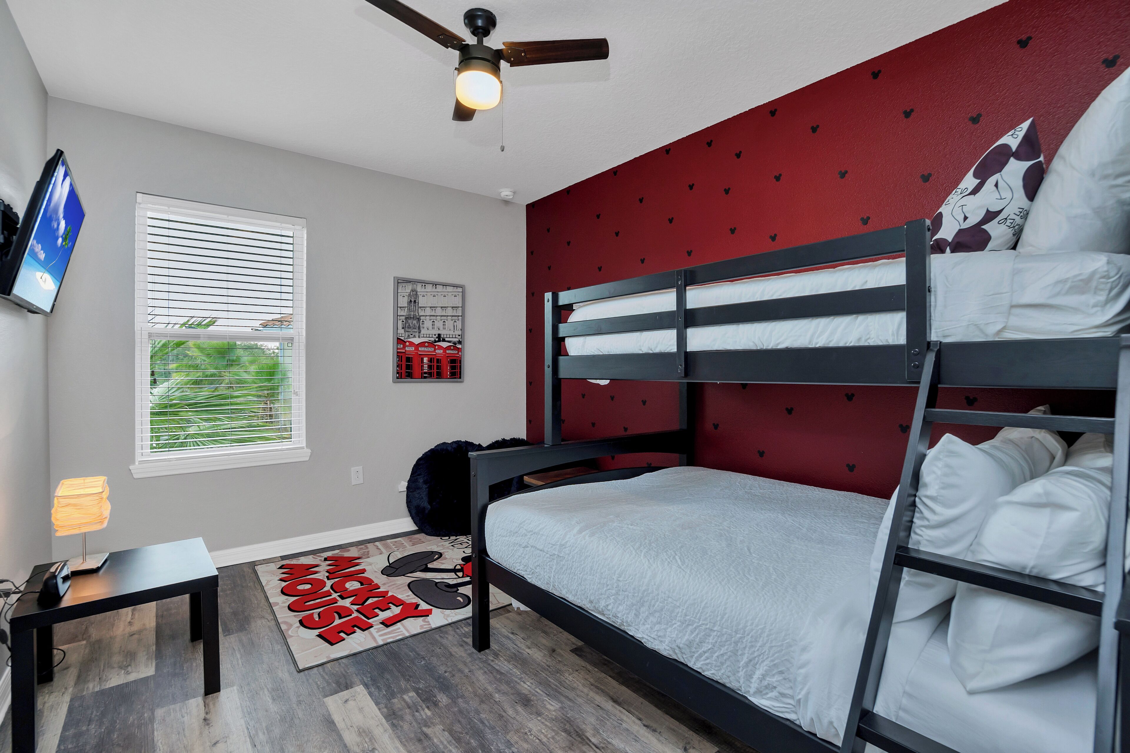 Habitación con literas, pared roja, ventilador de techo, ventana con persianas, mesita con lámpara y alfombra con temática de Mickey Mouse.