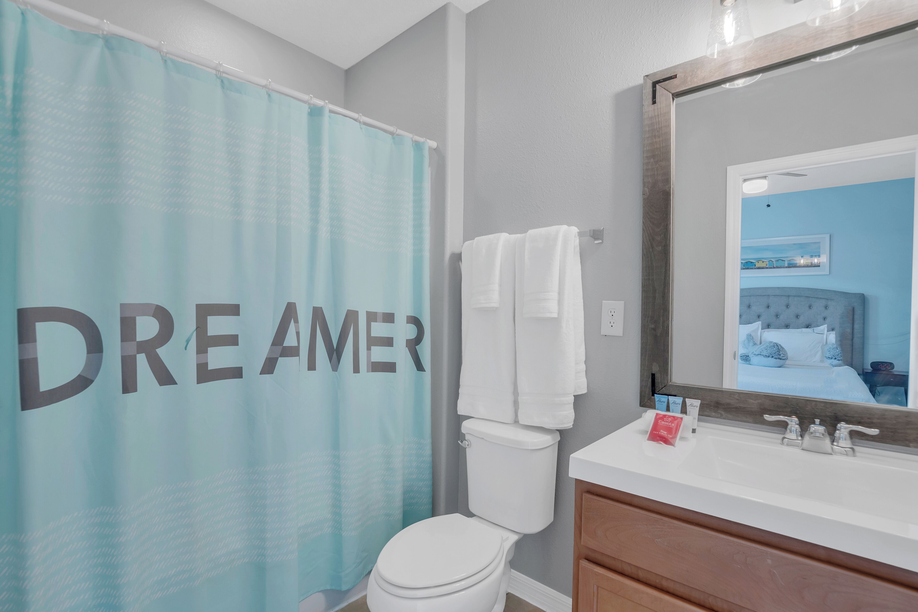 Une salle de bain avec un rideau de douche sur lequel est écrit le mot « RÊVEUR », des toilettes blanches, un meuble-lavabo avec un miroir et un lit visible dans le reflet.