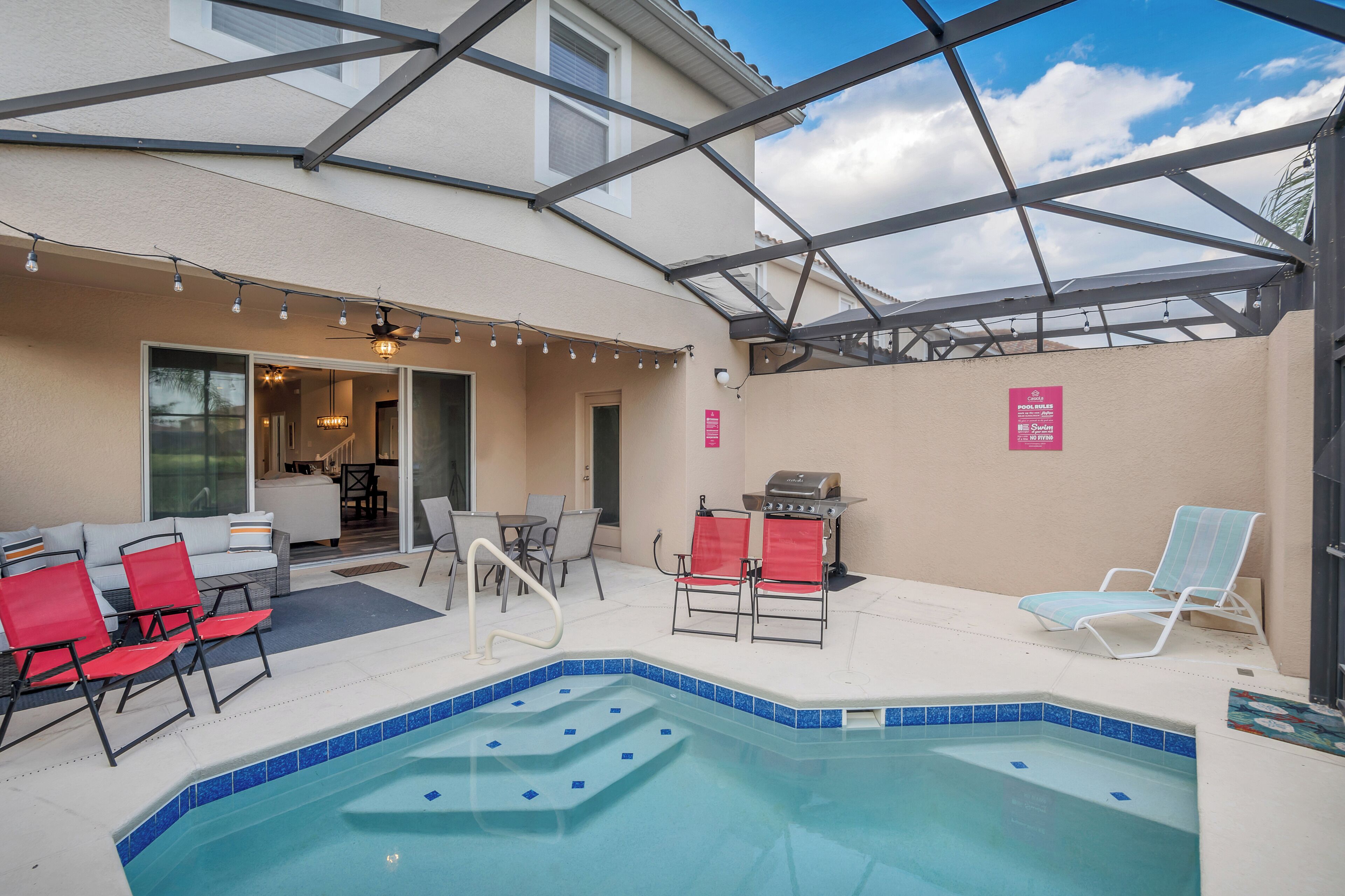 An outdoor pool area with a glass-enclosed patio, red and white lounge chairs, a barbecue grill, and a sign indicating a swimming area.