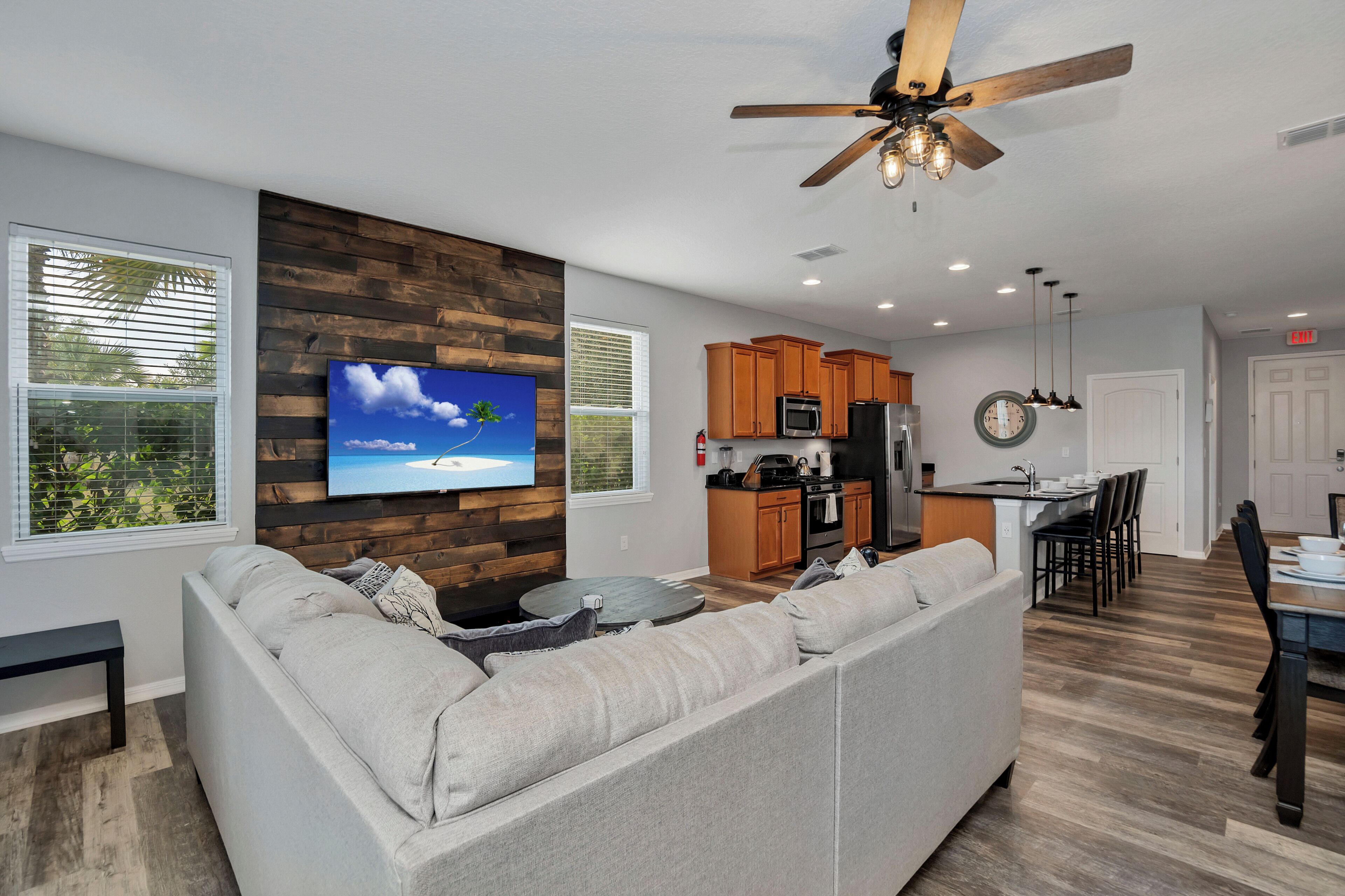 A modern living room with a gray sectional sofa, a flat-screen TV mounted on a wooden wall, a ceiling fan, and a dining area with a wooden table and chairs.