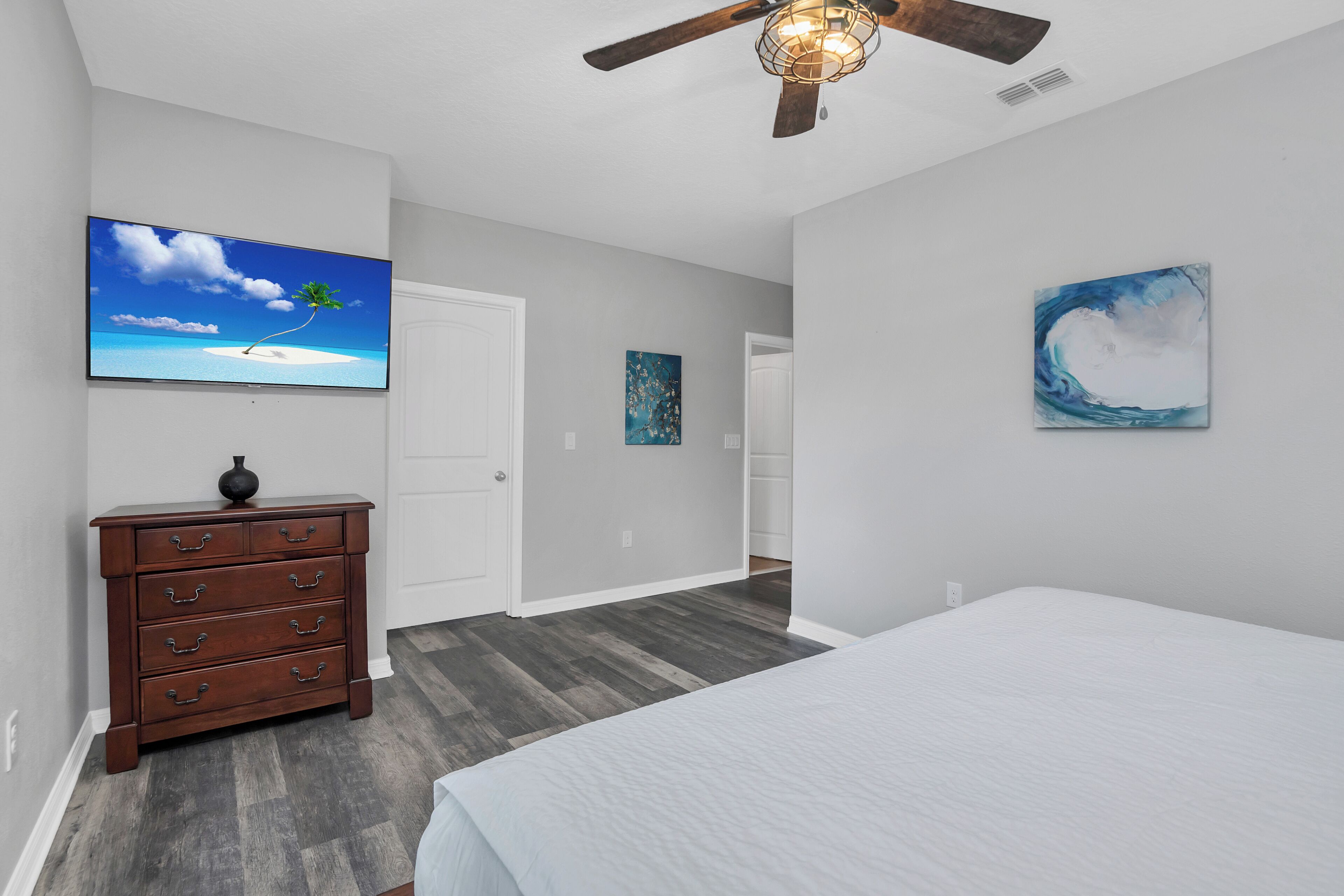 A bedroom with a bed, a dresser, a ceiling fan, and artwork on the walls.