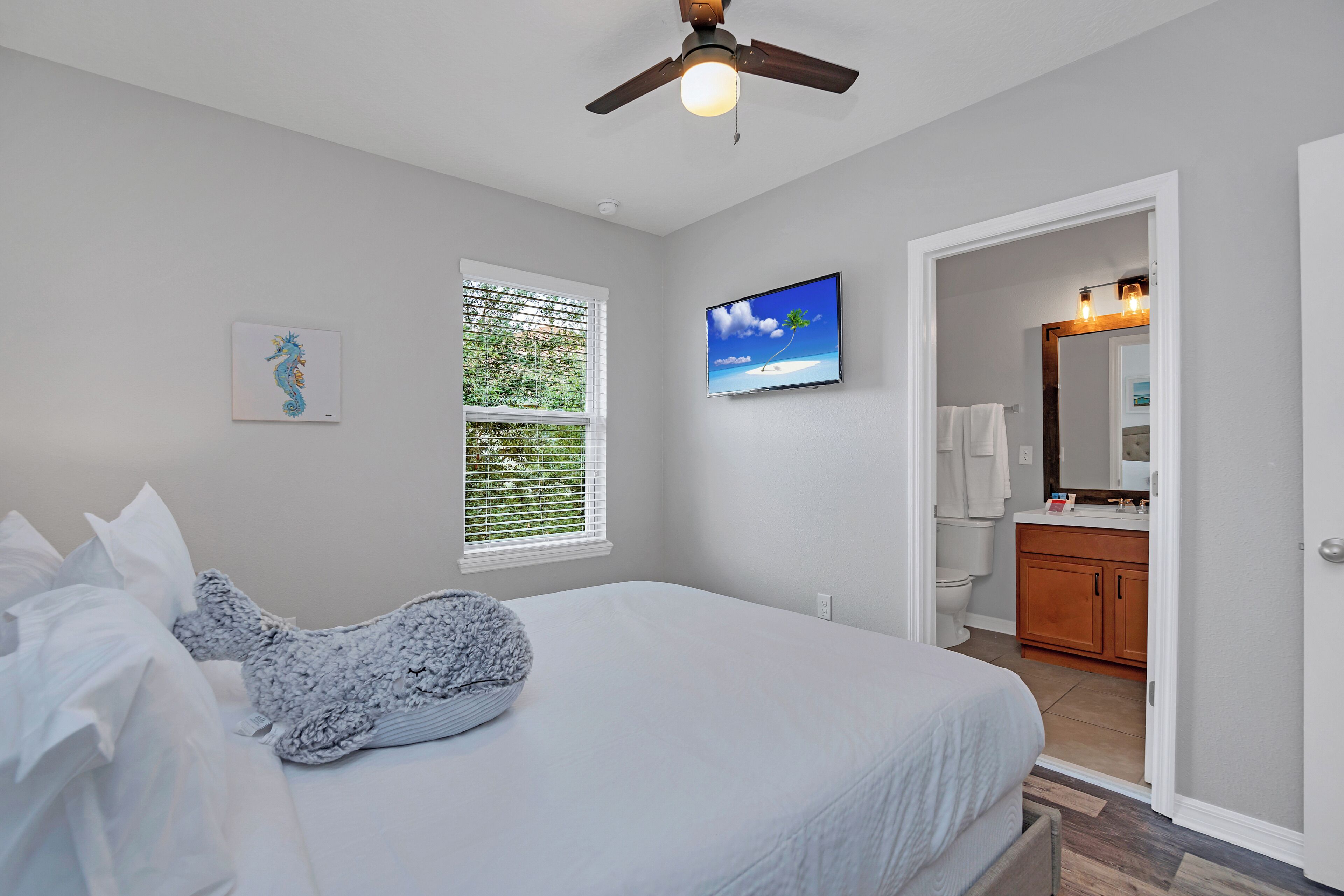 A bedroom with a bed, a ceiling fan, a window with blinds, a TV on the wall, and a bathroom visible through an open door.