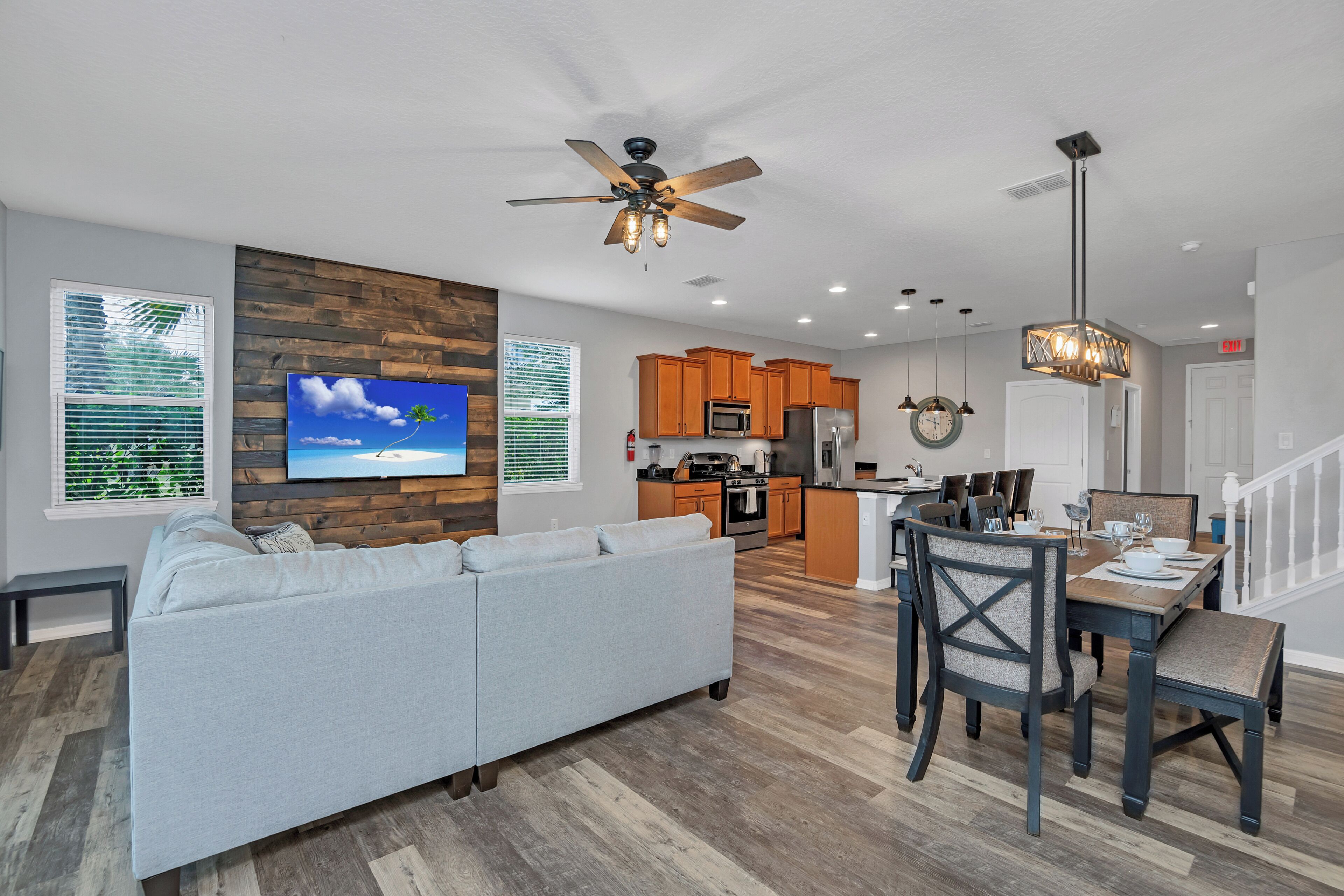 A modern living room with a gray sofa, wooden flooring, a ceiling fan, and a TV mounted on the wall.