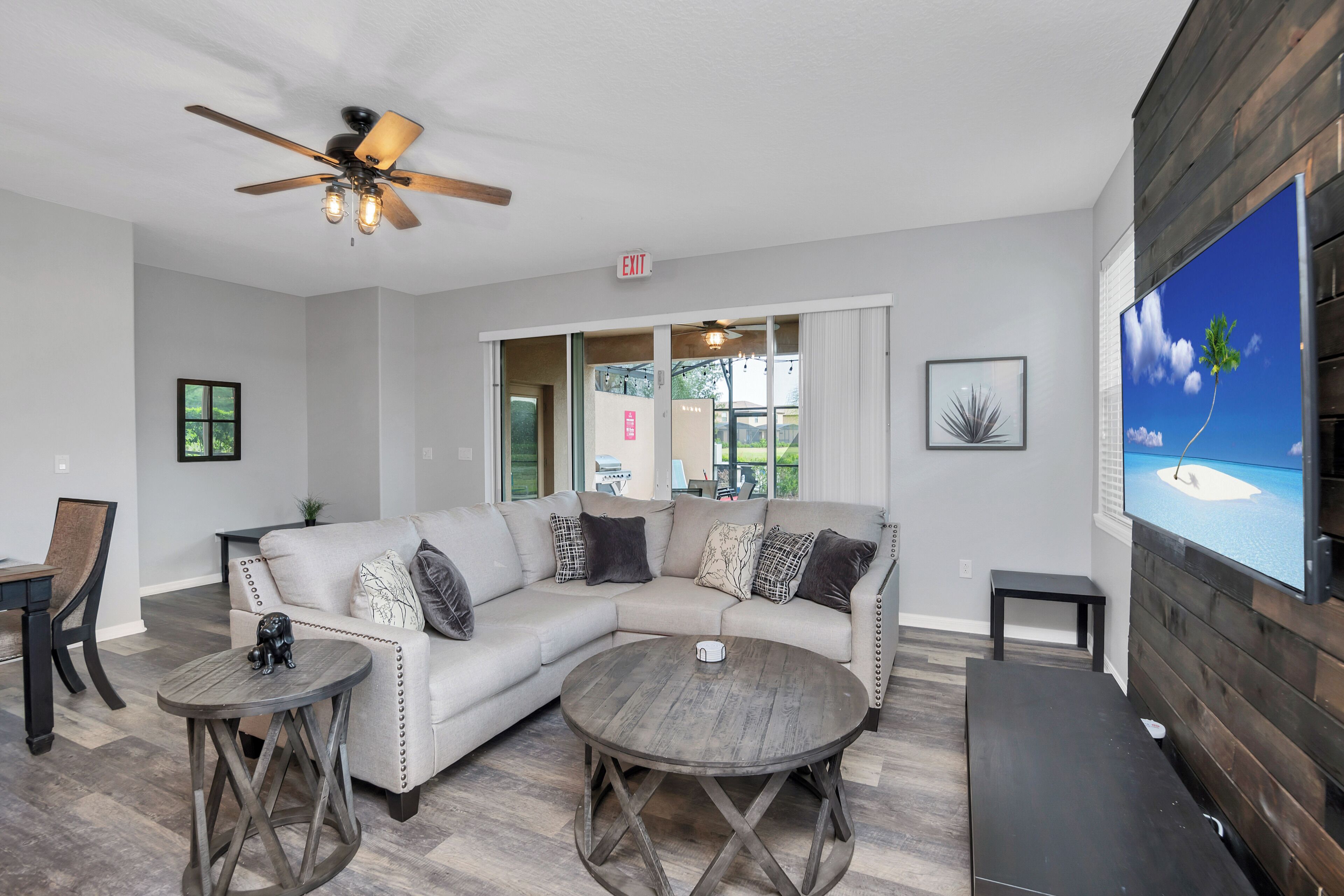 A modern living room with a sectional sofa, two round tables, a ceiling fan, and a large TV screen displaying a tropical scene.