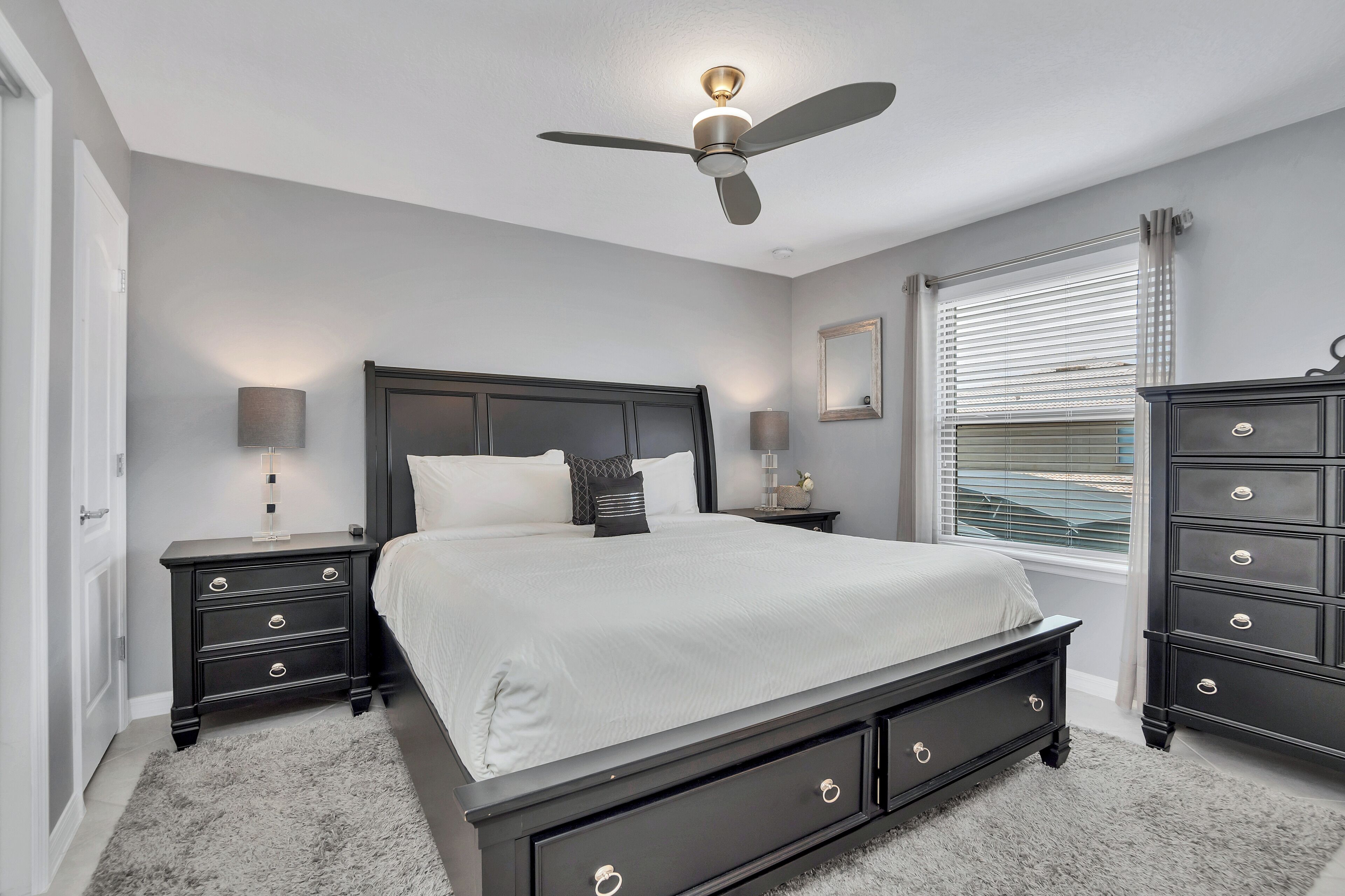 A bedroom with a large bed, two bedside tables, a ceiling fan, a window with blinds, and a dresser.