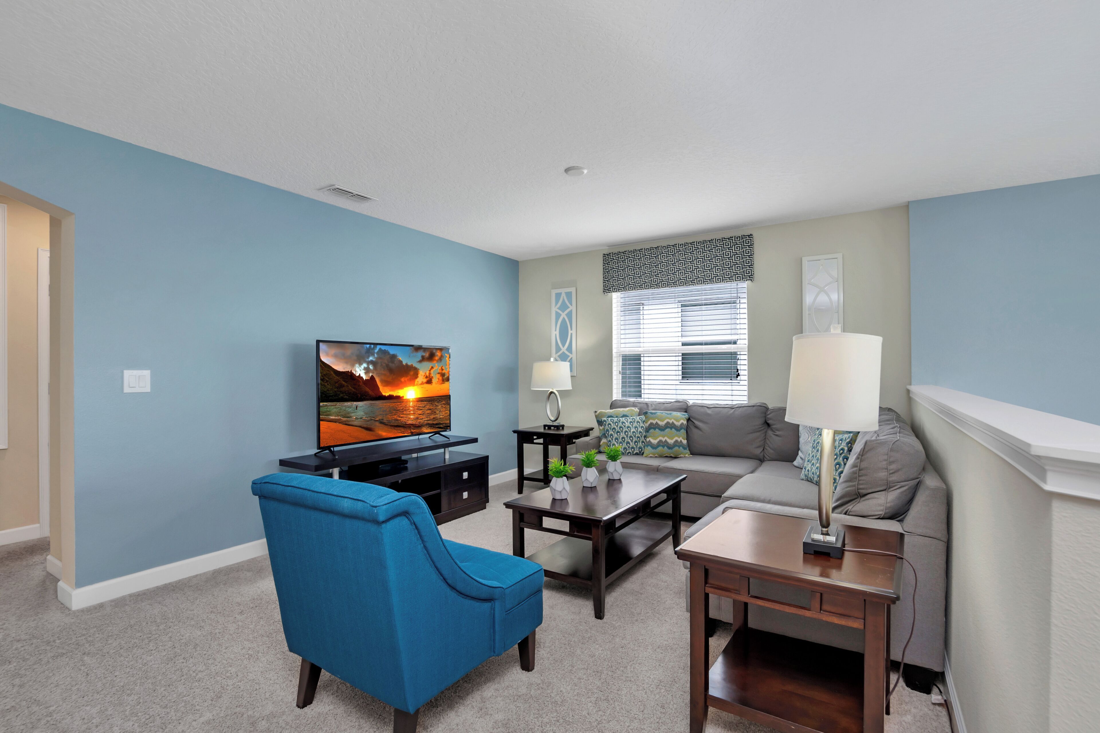 A living room with a blue armchair, a sofa, a coffee table, and a television displaying a sunset scene.