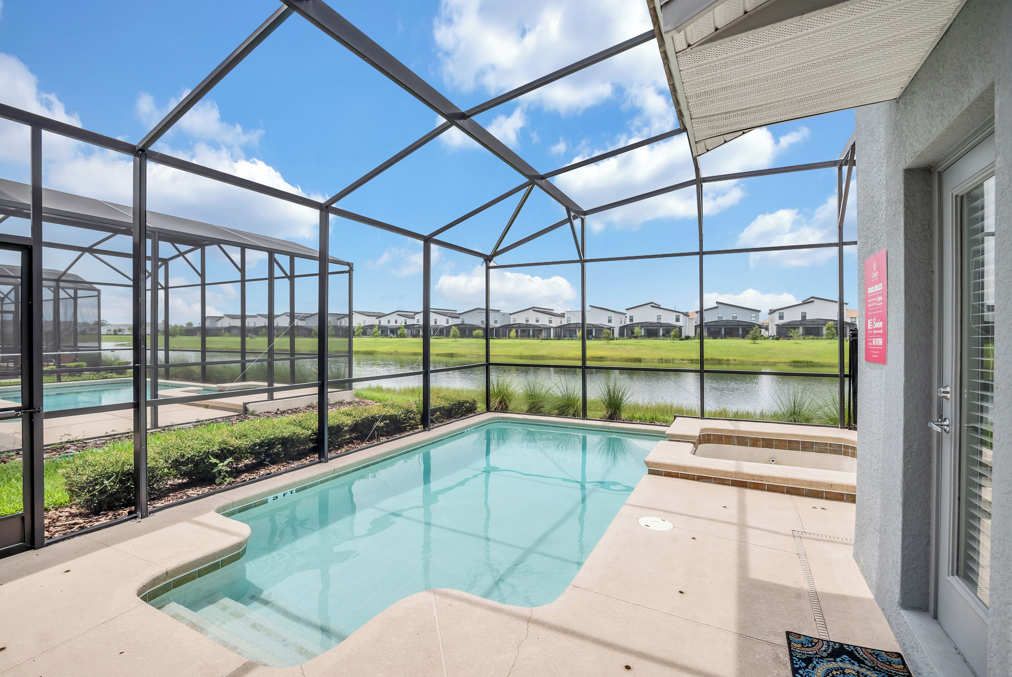 A pool area with a glass enclosure, a patio, and a view of a lake and houses in the background.
