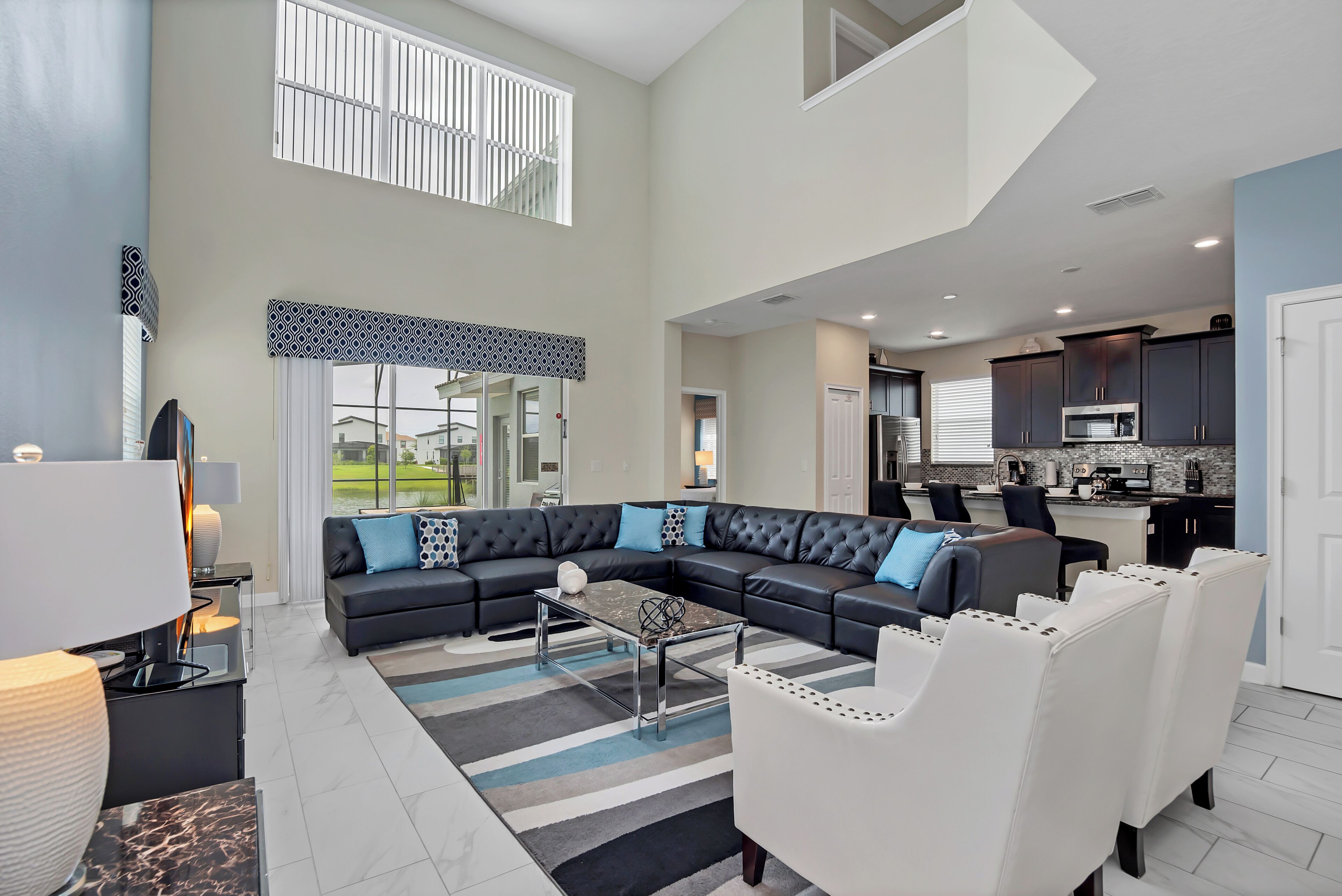 A spacious living room with a sectional sofa, a coffee table, and a dining area with white chairs. A kitchen is visible in the background.