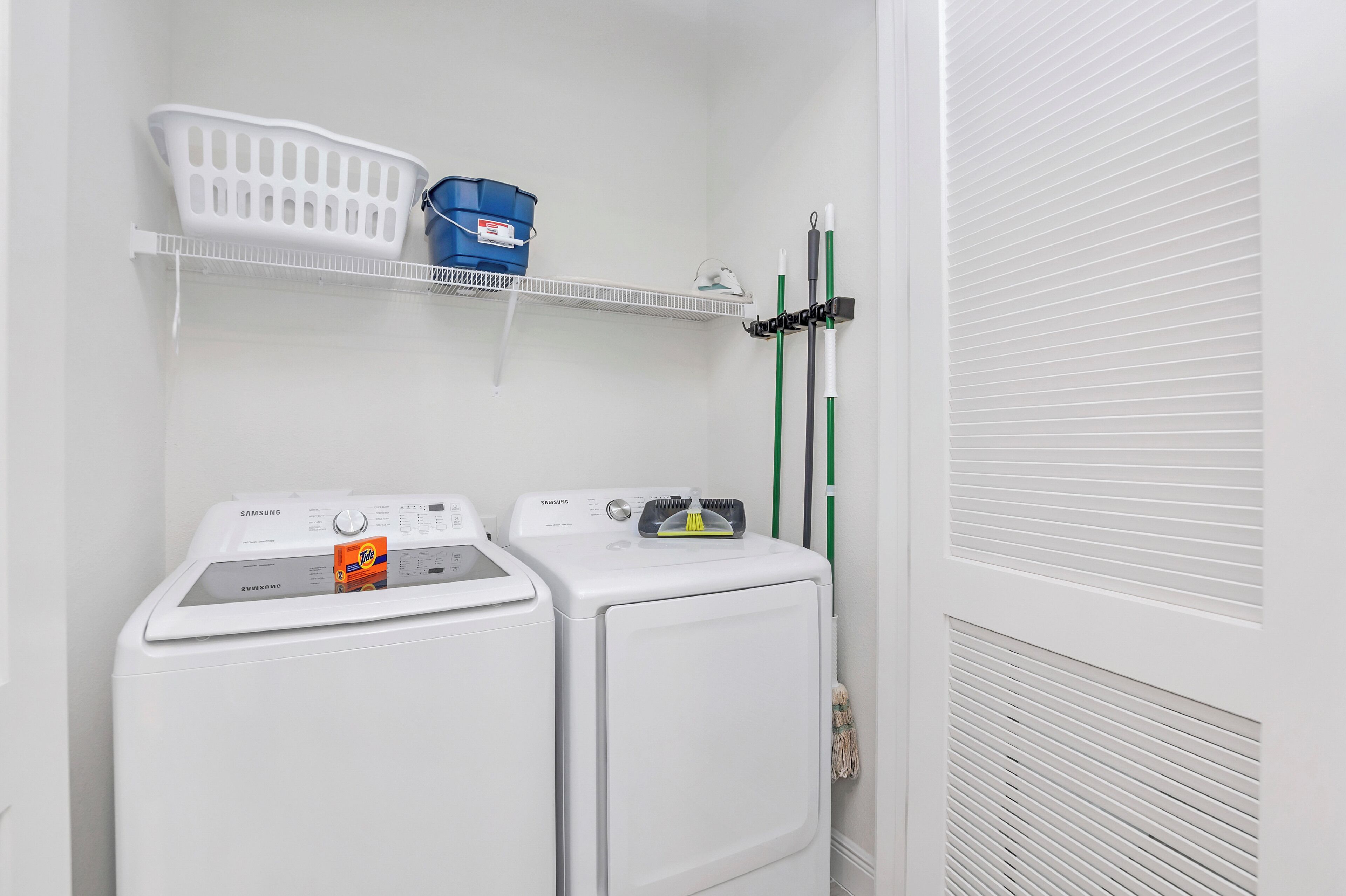 A laundry area with two white washing machines and a shelf above them holding a white basket and a blue bucket.