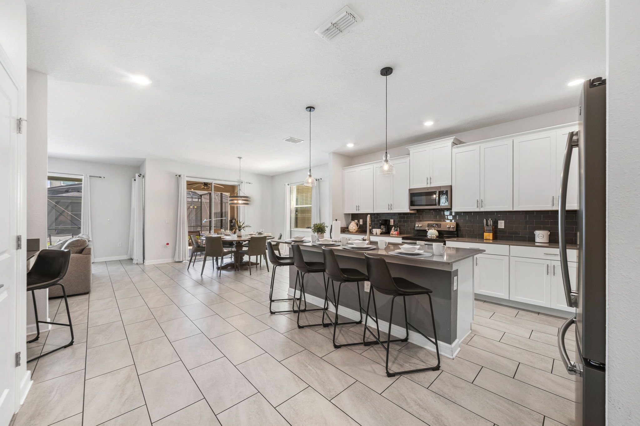A modern kitchen with white cabinets, a central island with bar stools, a dining area with chairs, and a visible refrigerator.