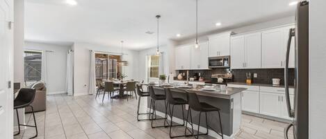 A modern kitchen with white cabinets, a central island with bar stools, a dining area with chairs, and a visible refrigerator.
