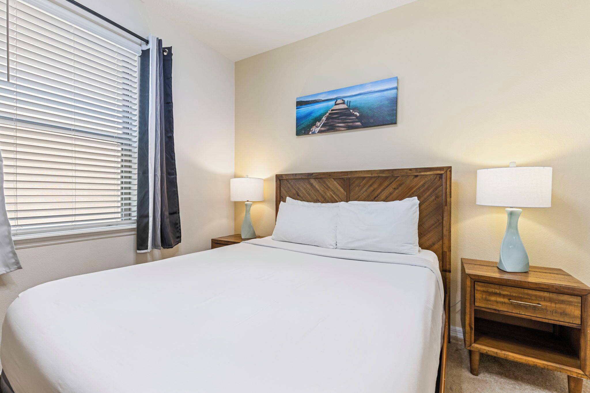 A neatly made bed with white linens, two bedside tables with lamps, a wooden headboard, a window with blinds and a curtain, and a painting of a pier on the wall.