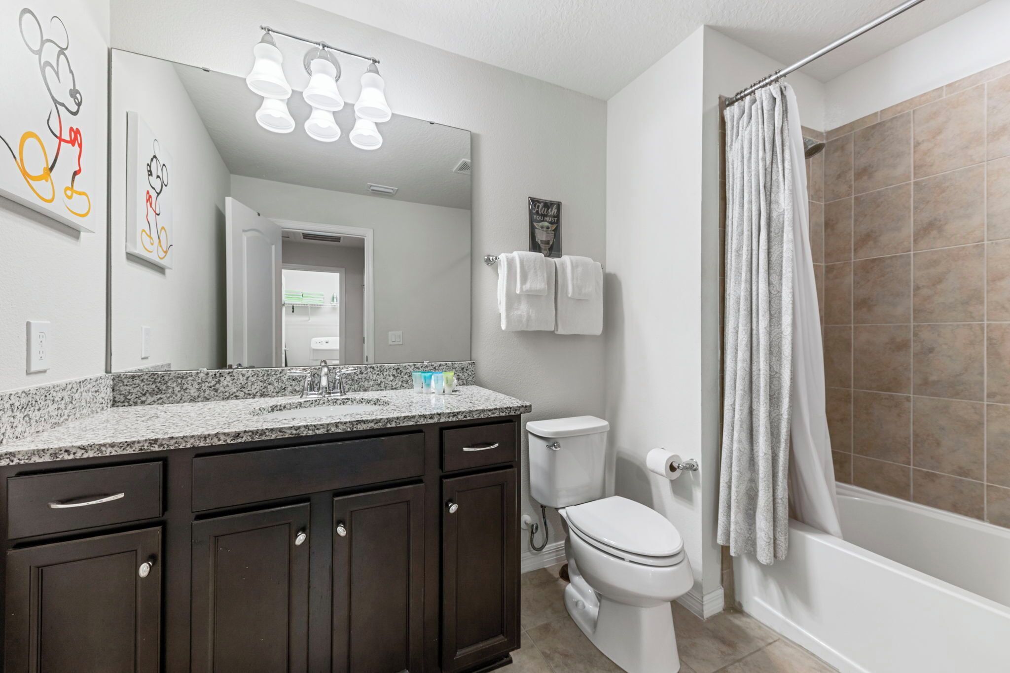 A modern bathroom with a granite countertop, dark wooden cabinets, a white toilet, a shower with a beige tiled wall, and a large mirror with wall art.