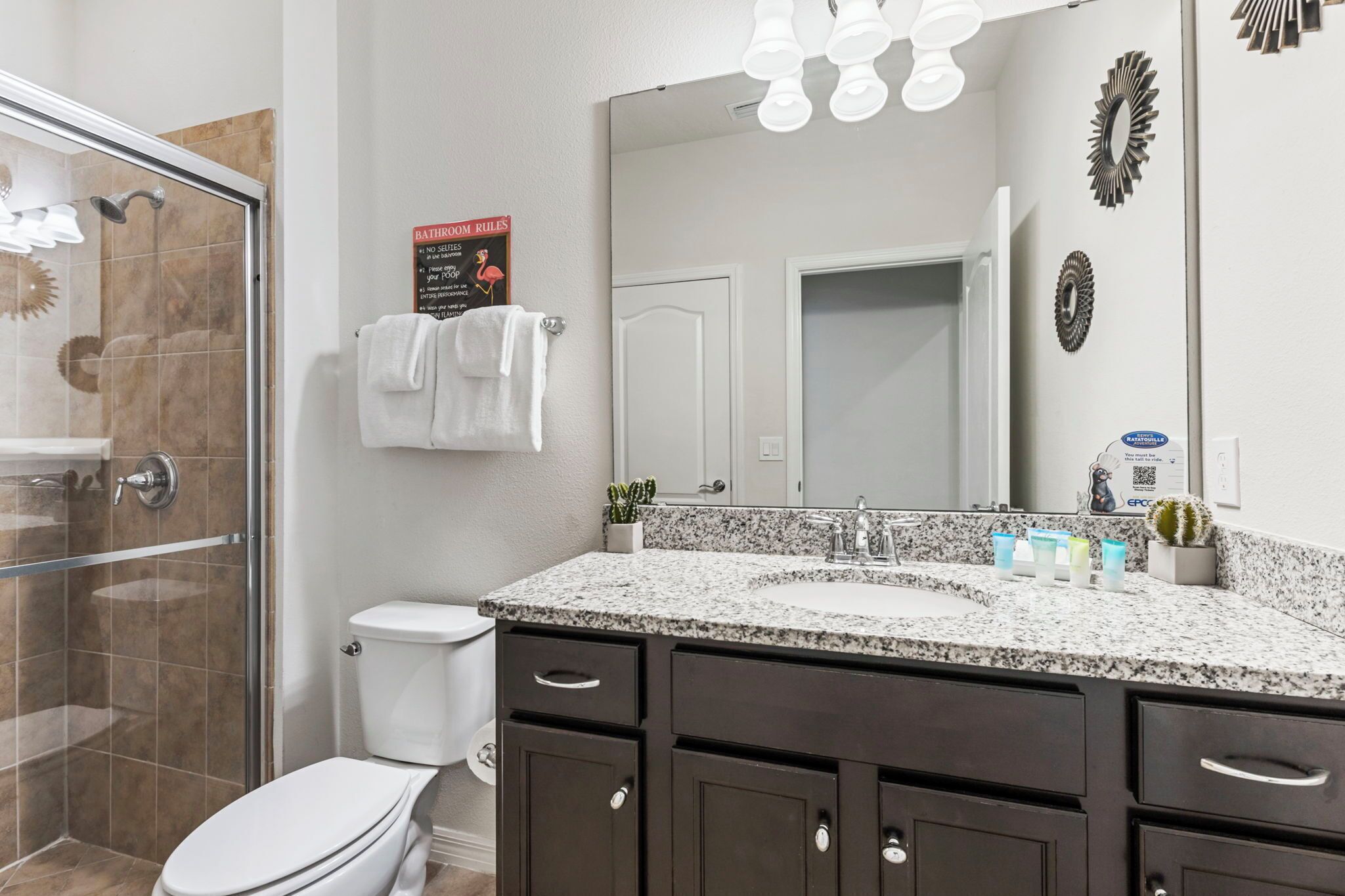A bathroom with a glass shower enclosure, a white toilet, dark cabinets, a granite countertop, and a large mirror.