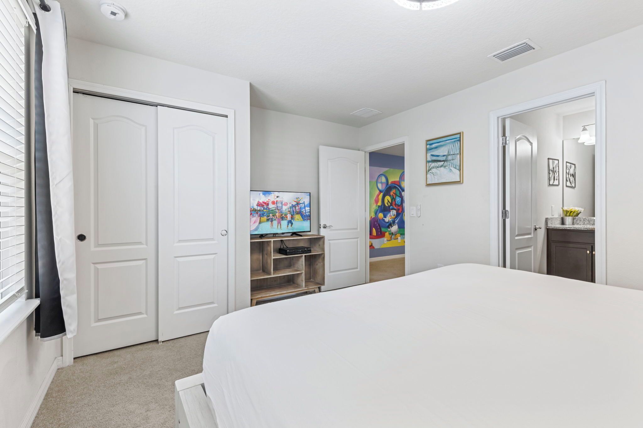 A hotel room with a large bed, a television on a stand, a door with a colorful painting, and a bathroom with a mirror and sink.