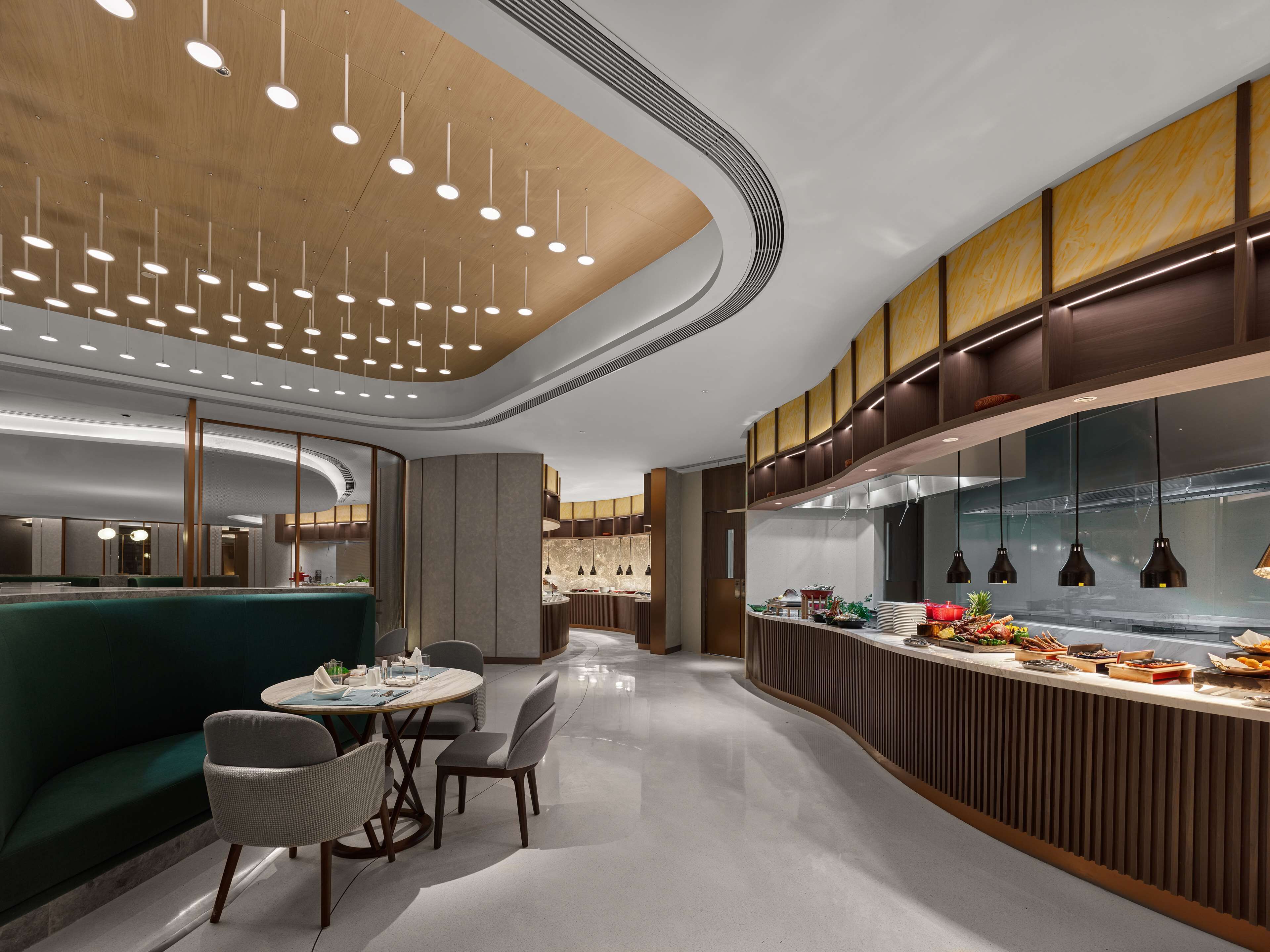 Un interior de restaurante moderno con una zona de comedor central, una barra con exhibición de alimentos y una cocina al fondo.