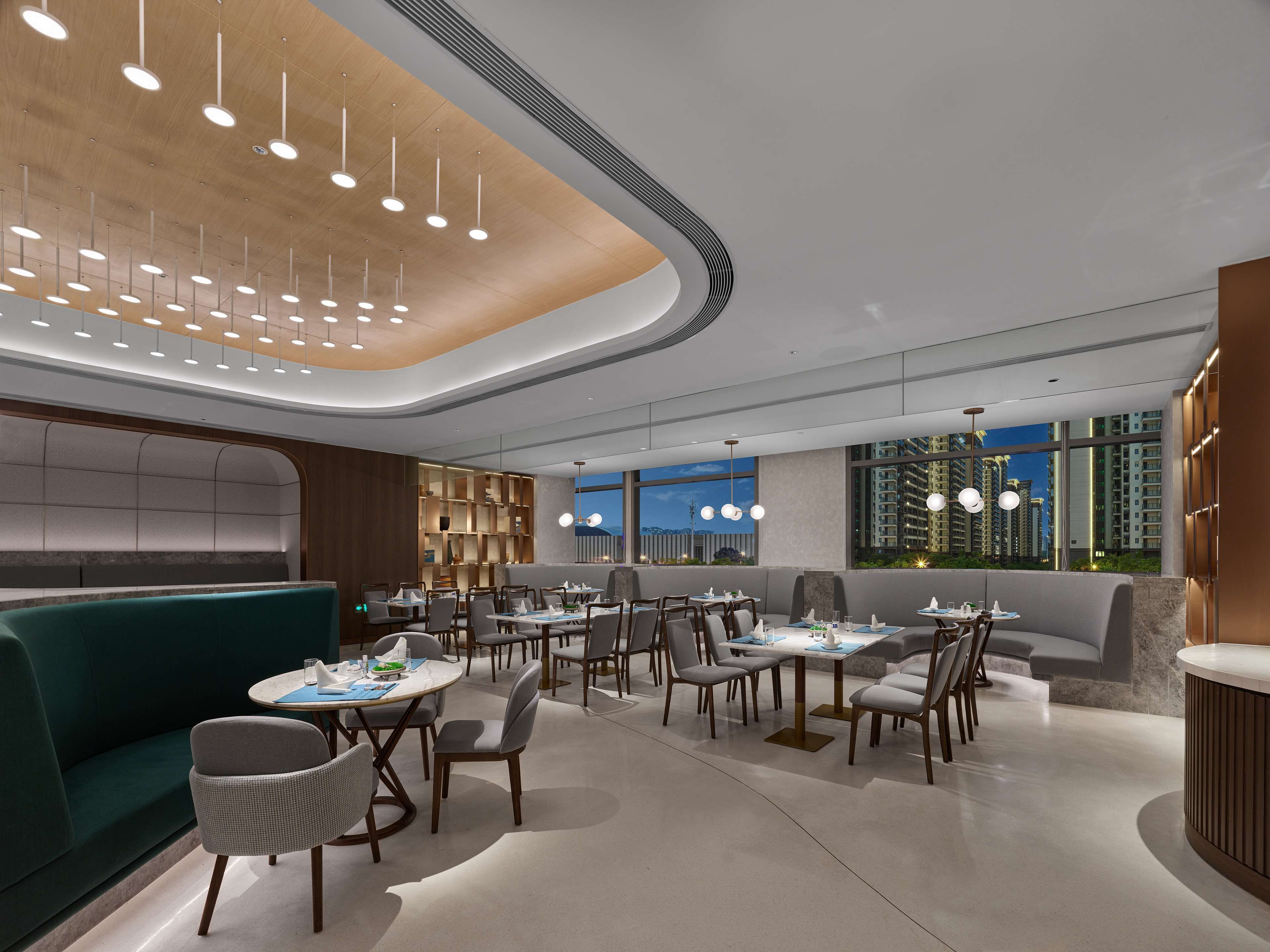 Ein modernes Restaurant mit runden Tischen, grünen Sitzgelegenheiten und Blick auf hohe Gebäude.