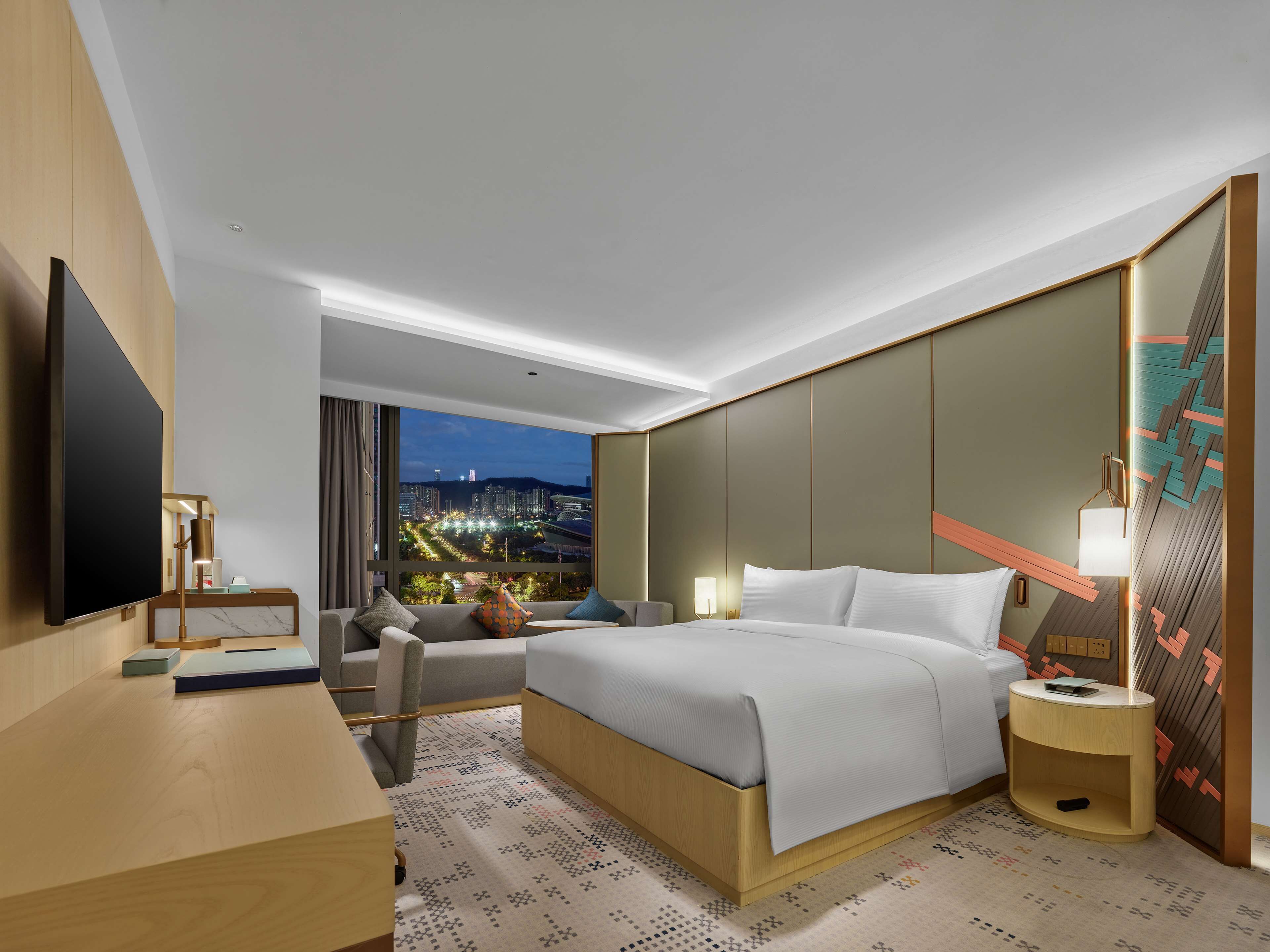 A modern hotel room with a large bed, a desk, a TV, and a view of the cityscape at night.