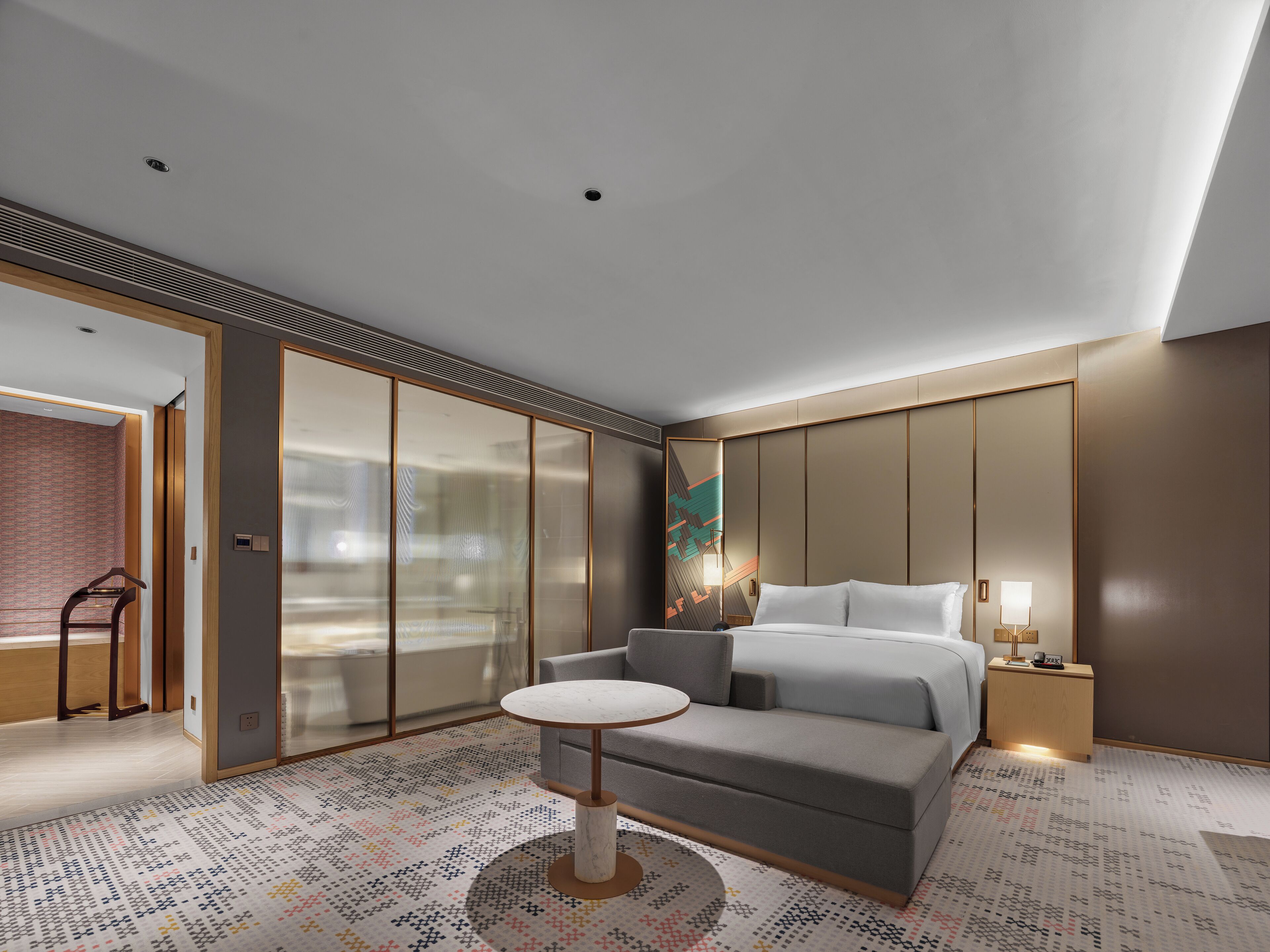 Una habitación de hotel moderna con un sofá gris, una mesita redonda pequeña, una cama con ropa blanca, una mesita de noche con lámpara y un espejo grande.
