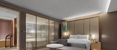 Una habitación de hotel moderna con un sofá gris, una mesita redonda pequeña, una cama con ropa blanca, una mesita de noche con lámpara y un espejo grande.