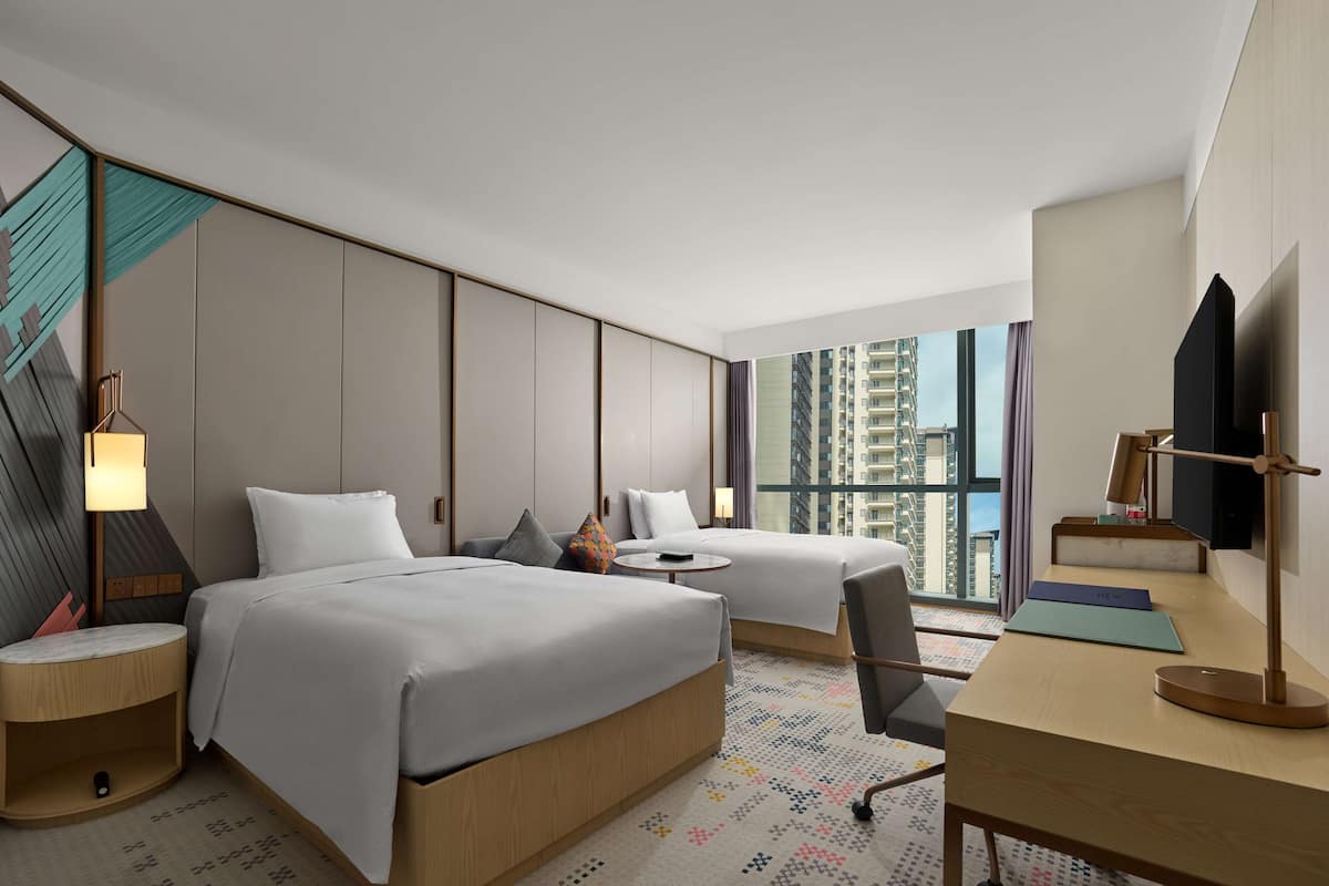 A modern hotel room with two beds, a desk, a chair, a TV, and a large window offering a city view.