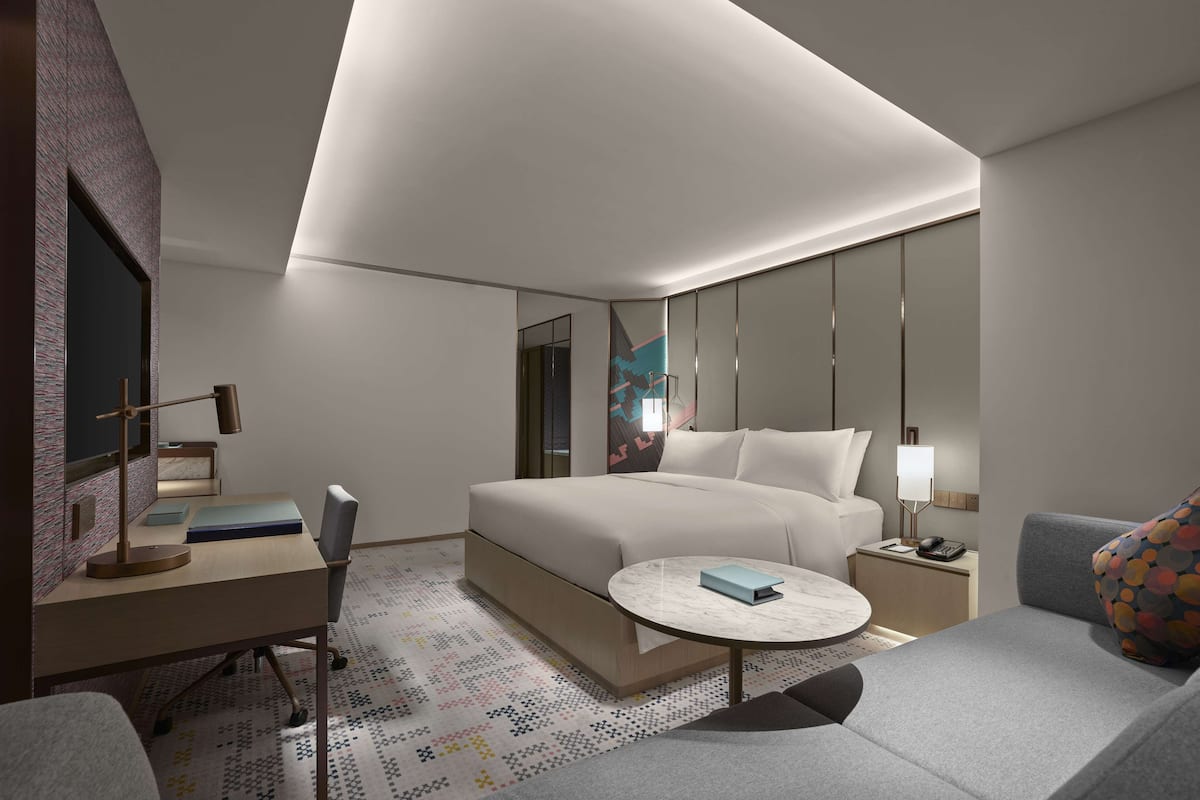 A modern hotel room with a large bed, a desk with a chair, a round coffee table, and a sofa.