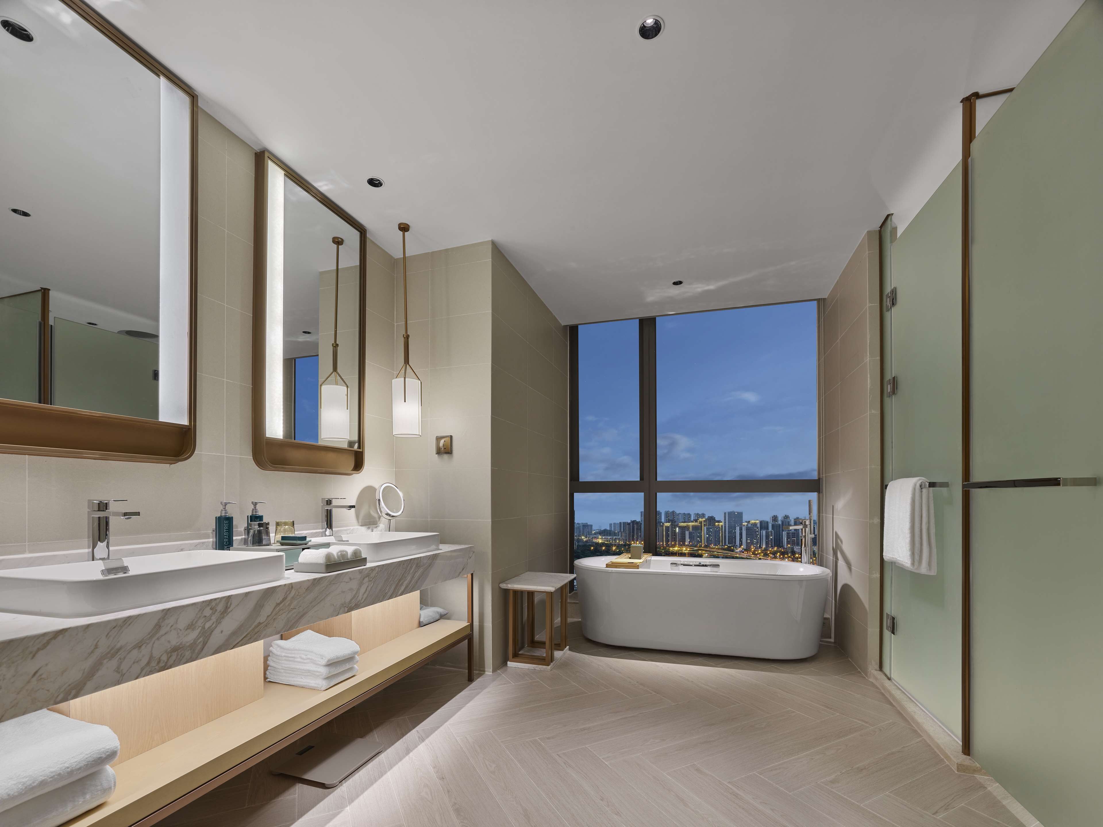 Un baño moderno con lavamanos dobles, un espejo grande, una bañera y vistas a la ciudad a través de un amplio ventanal.