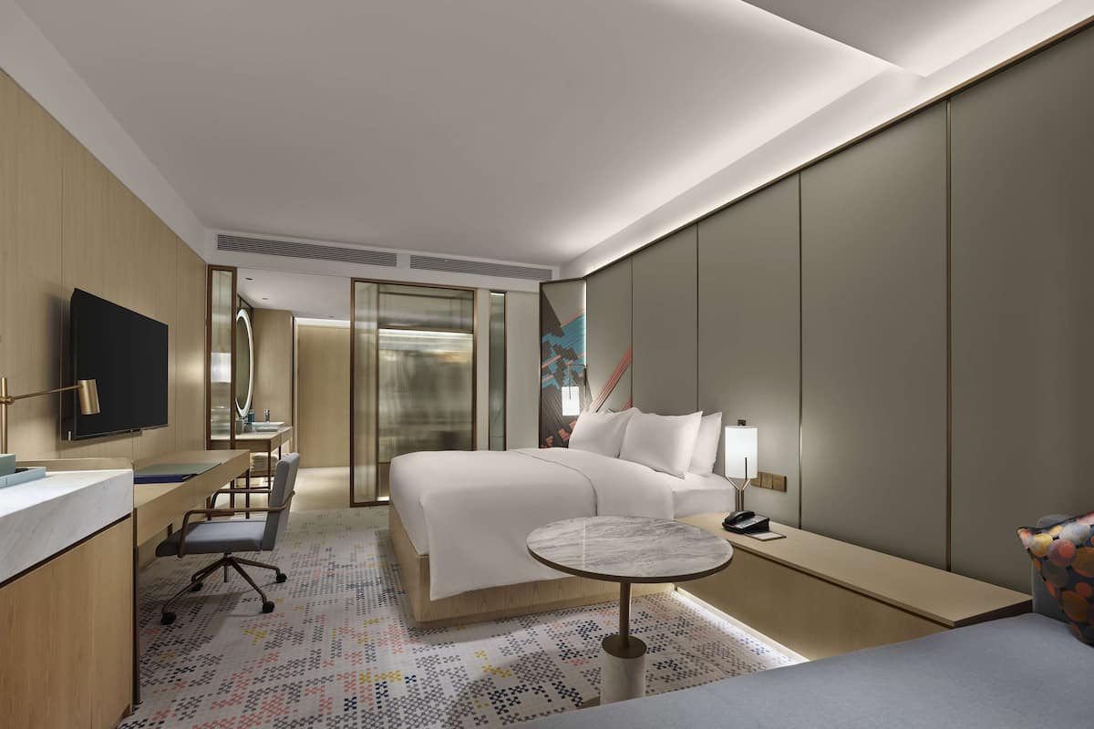A modern hotel room with a bed, bedside table, desk, chair, and a TV.