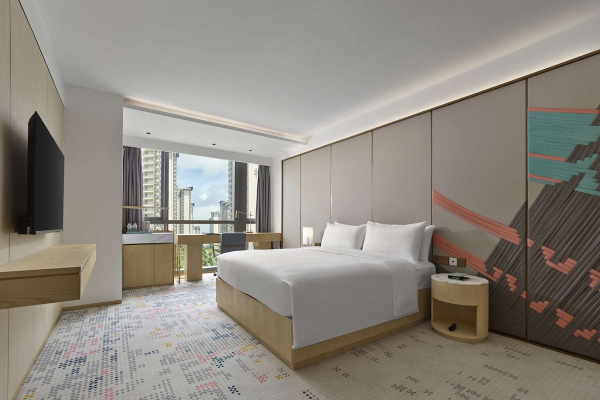 A modern hotel room with a large bed, bedside tables, a TV, and a city view through the window.