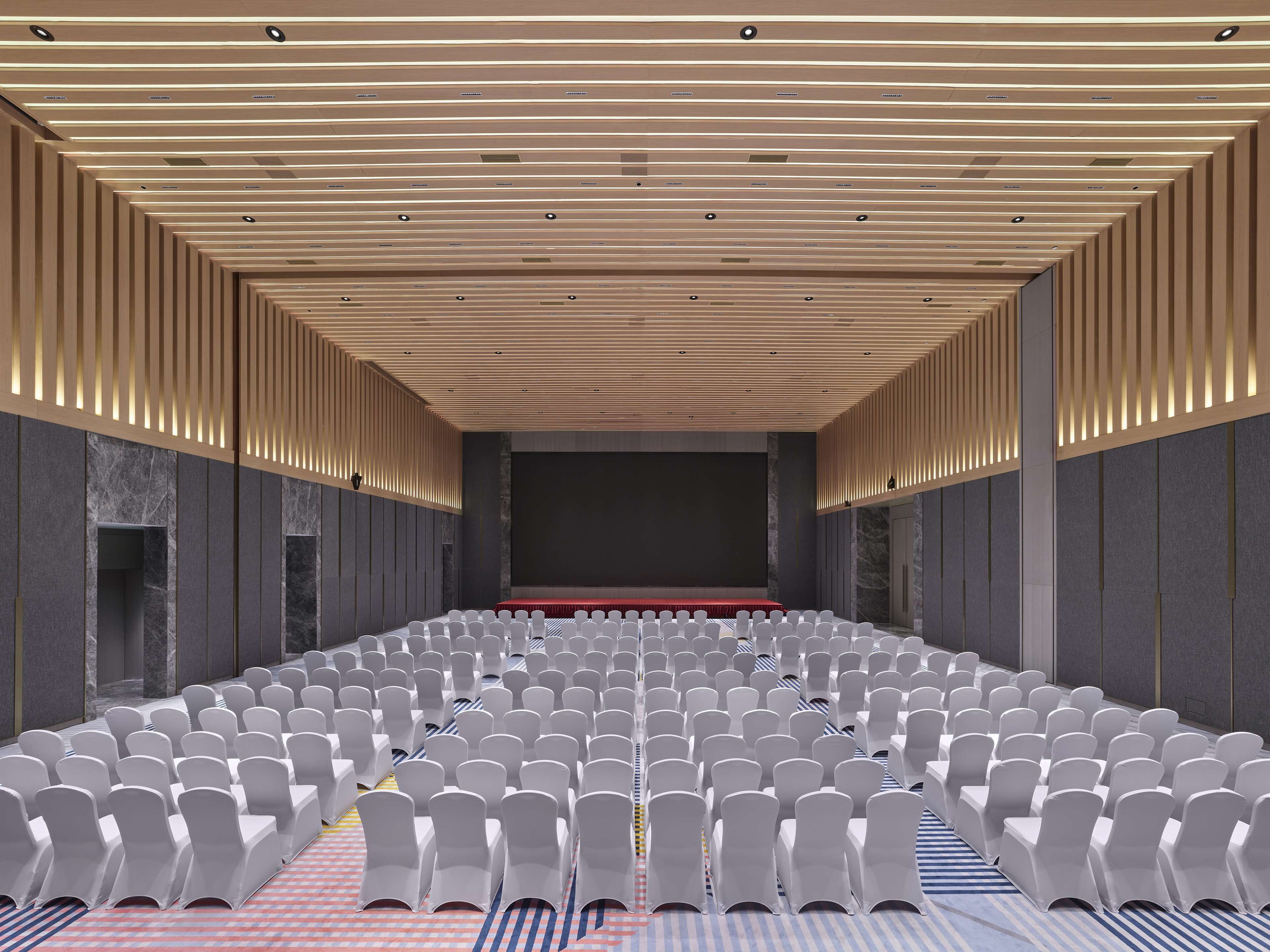 Une salle de conférence avec des rangées de chaises blanches, une scène et un plafond en bois à lattes.