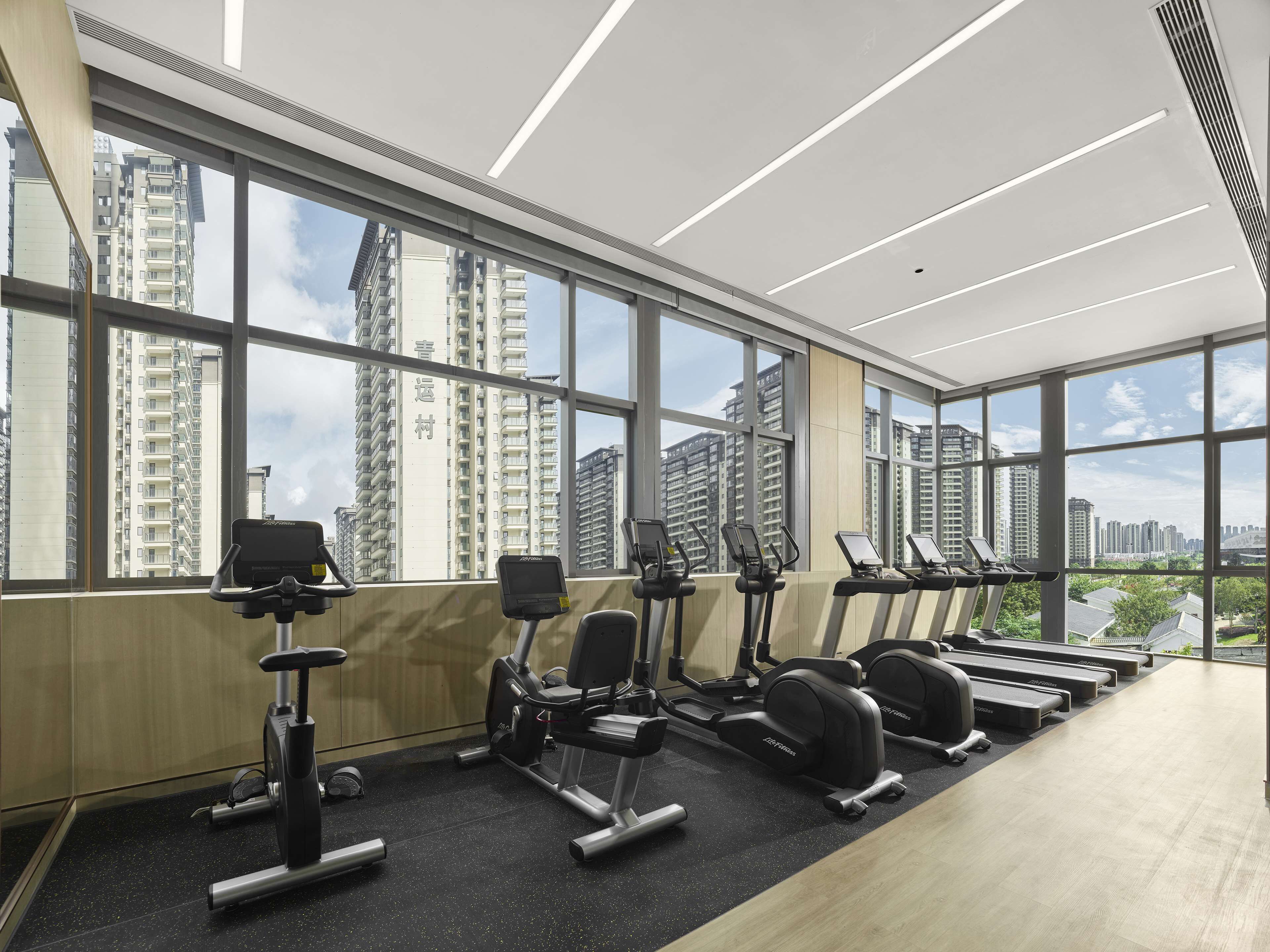 Ein Fitnessstudio mit Trainingsgeräten und großen Fenstern, die einen Blick auf hohe Gebäude ermöglichen.