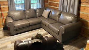 A brown leather sectional sofa with a cushion, a wooden floor, and a window with blinds. - Valentine Drive Cabin 1 (Lookout Mountain)
