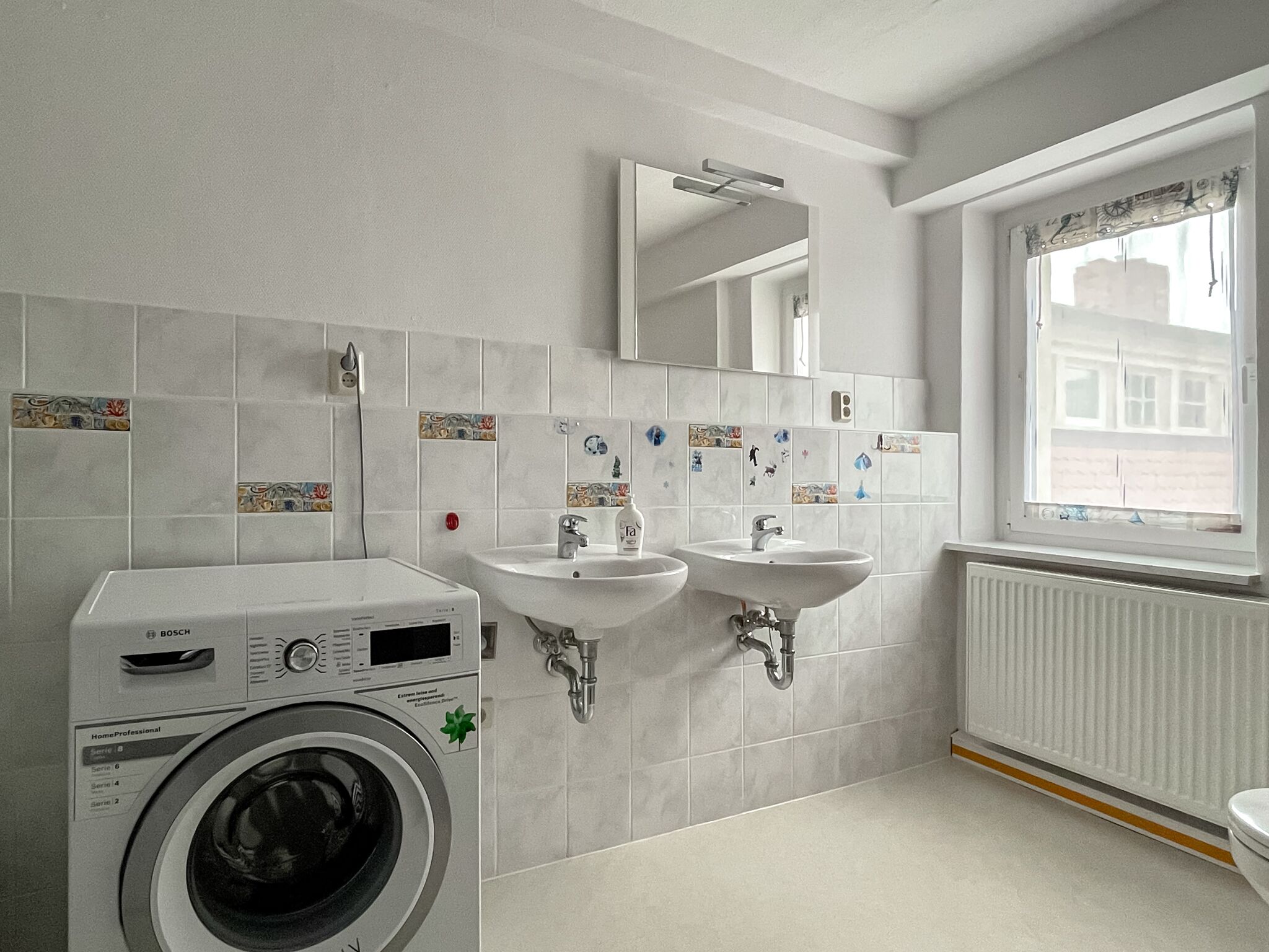 A modern bathroom with a Bosch washing machine, two sinks, a mirror, and a window with a view of buildings.