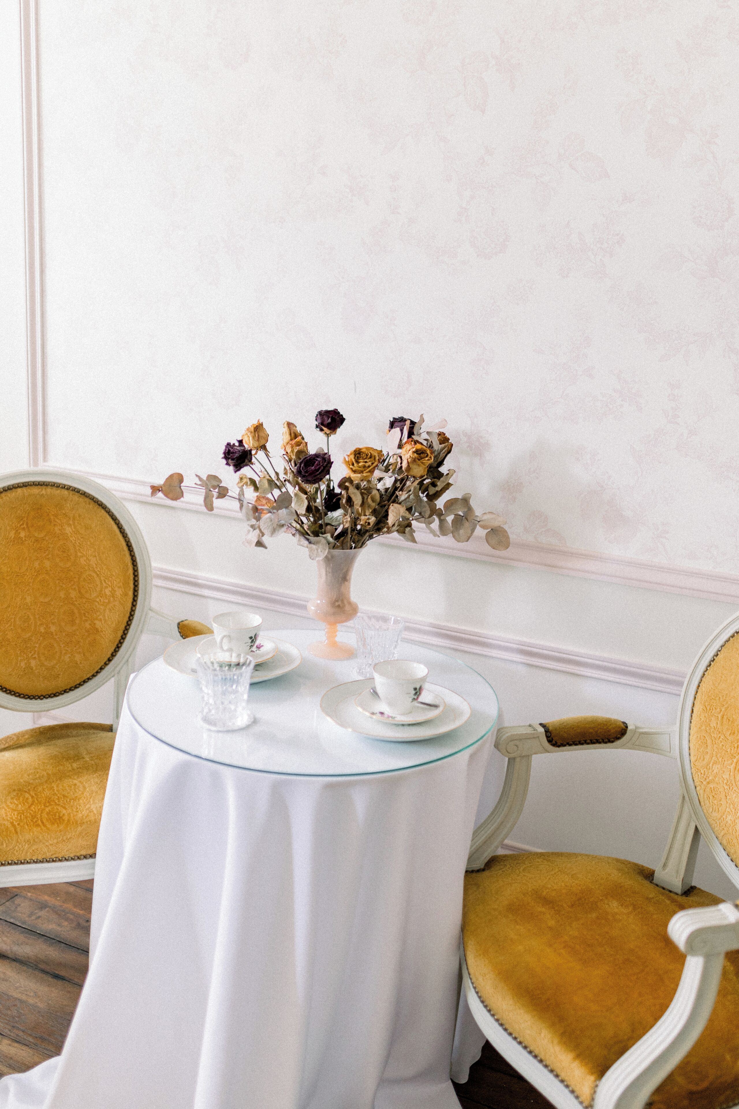 Ein Tisch mit weißer Tischdecke, einer Vase mit getrockneten Blumen, einer Teetasse und einem Untertasse.