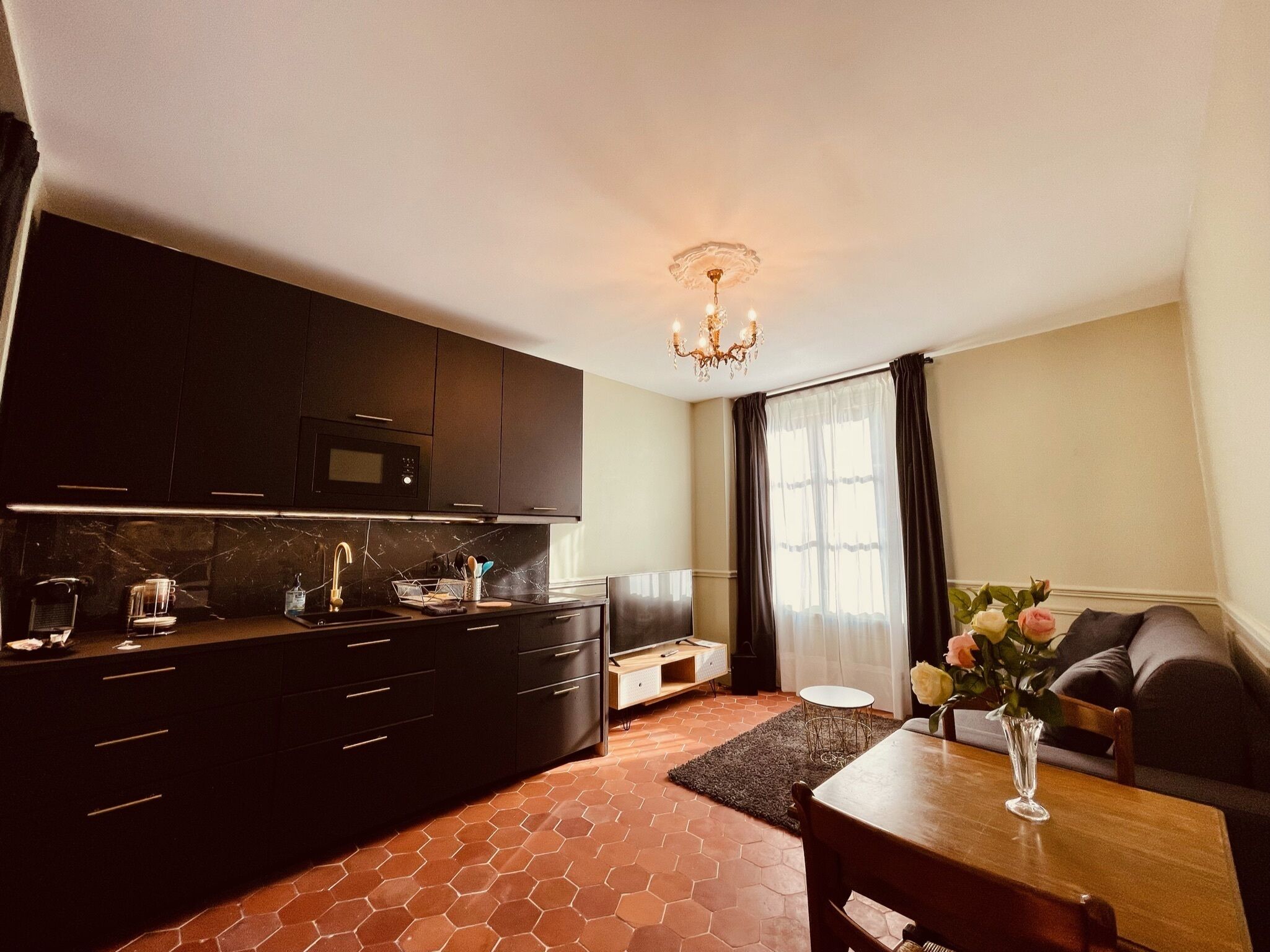 A compact kitchen with dark cabinetry, a microwave, and a sink. There's a dining area with a table and chairs, and a sofa. A chandelier hangs from the ceiling.
