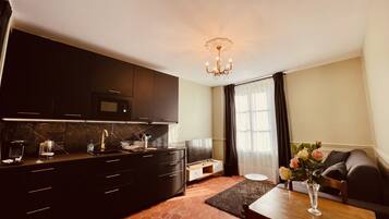 Eine kleine Küche mit dunklen Schränken, einer Mikrowelle und einem Spülbecken. Es gibt einen Essbereich mit Tisch und Stühlen, sowie ein Sofa. Eine Pendelleuchte hängt von der Decke.