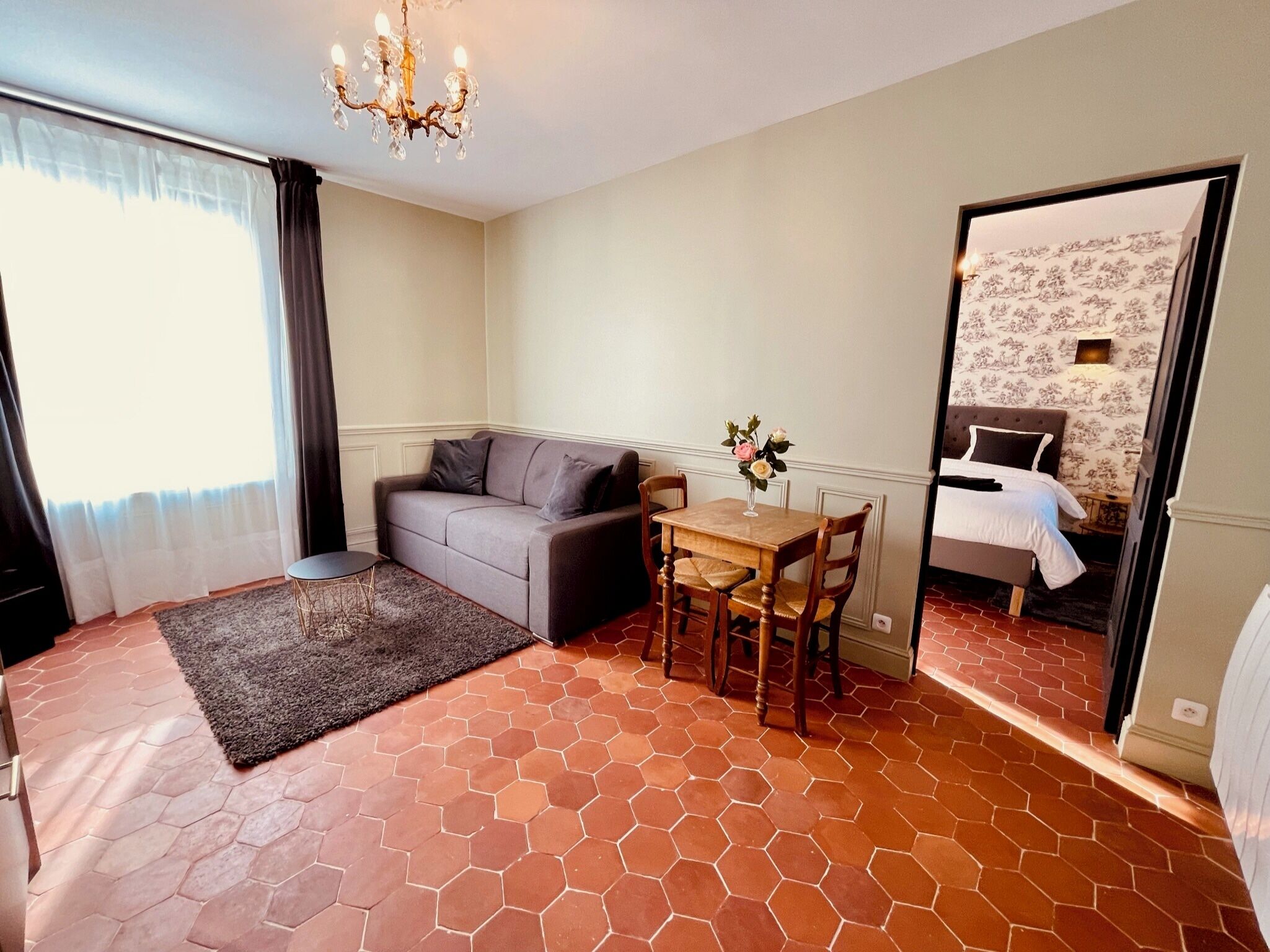 Una habitación de hotel con un sofá gris, una mesita con un florero y una cama con papel tapiz floral.