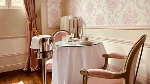 Un comedor con una mesa redonda preparada para dos, una silla, una lámpara y una ventana con cortinas.