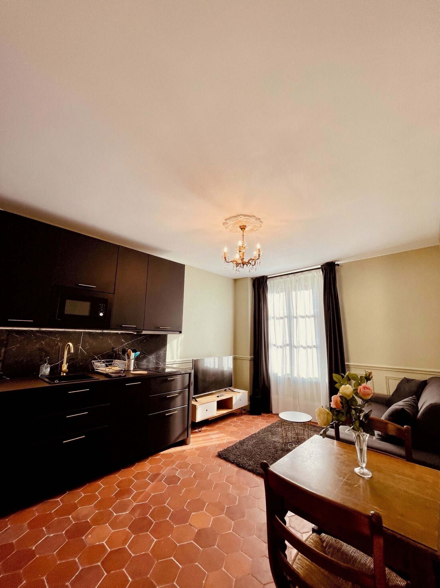 A modern kitchen with dark cabinetry, a microwave, and a TV. There's a dining area with a table and chairs, and a chandelier above.