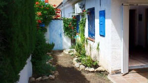 Un étroit sentier mène à un bâtiment blanc aux volets bleus, devant lequel se trouve un jardin parsemé de fleurs rouges.