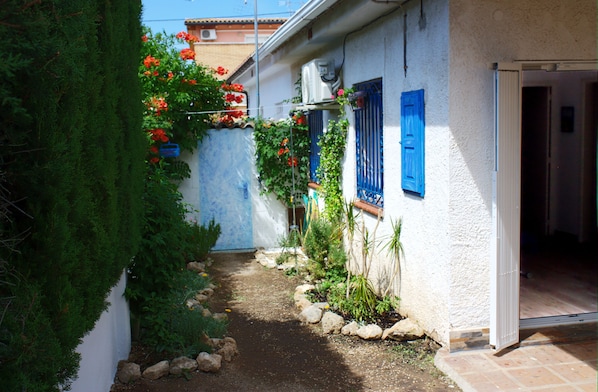 A narrow pathway leading to a white building with blue shutters and a garden with red flowers.