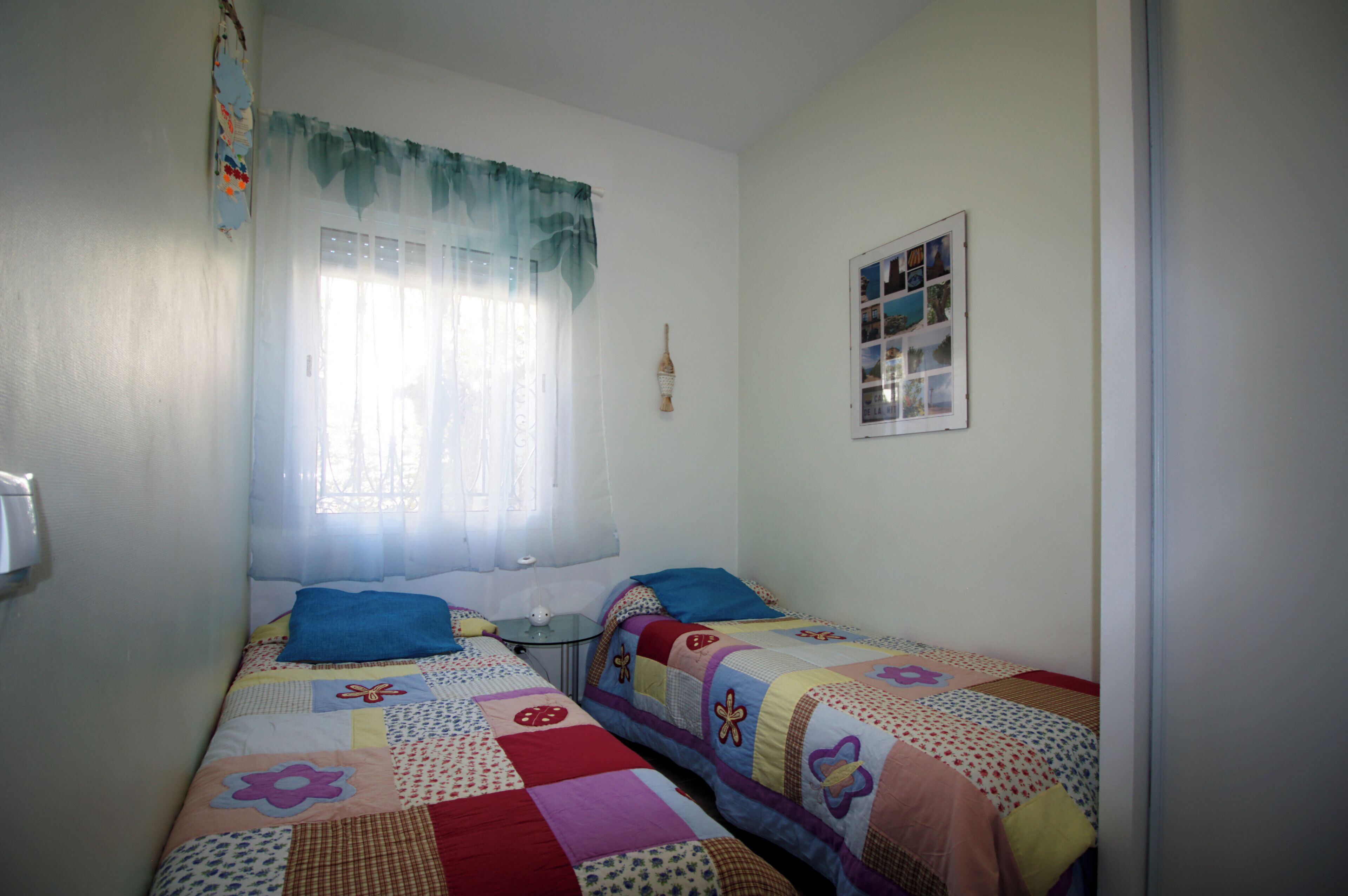 A small, well-lit bedroom with two single beds, a bedside table, a window with sheer curtains, and a wall-mounted photo collage.